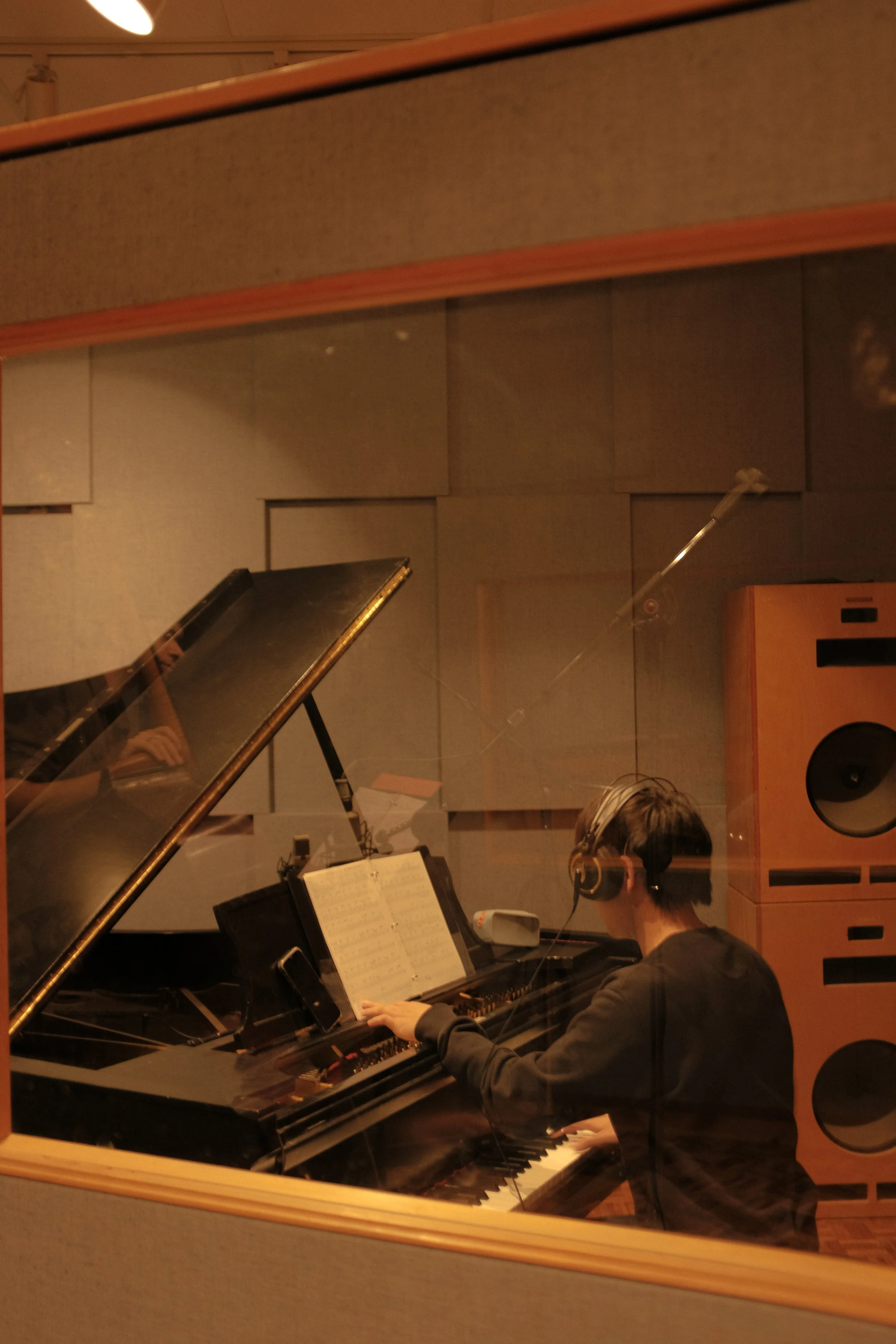 A person playing a grand piano in a recording studio, seen through a glass window with a reflection of a person's hand and part of their arm.