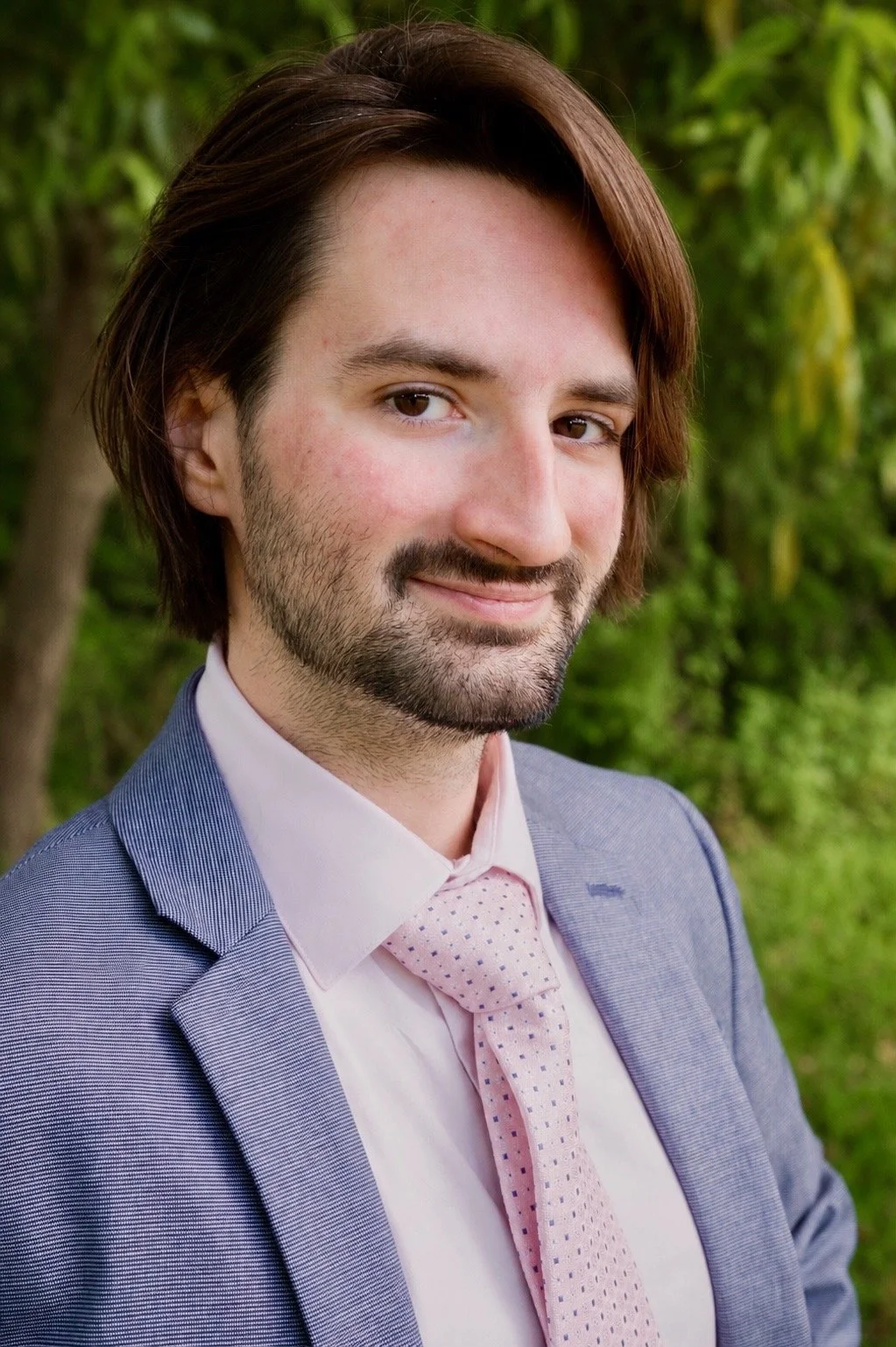 A man with brown hair and a beard, dressed in a light blue suit with a white shirt and pink tie, smiling outdoors with green trees in the background.