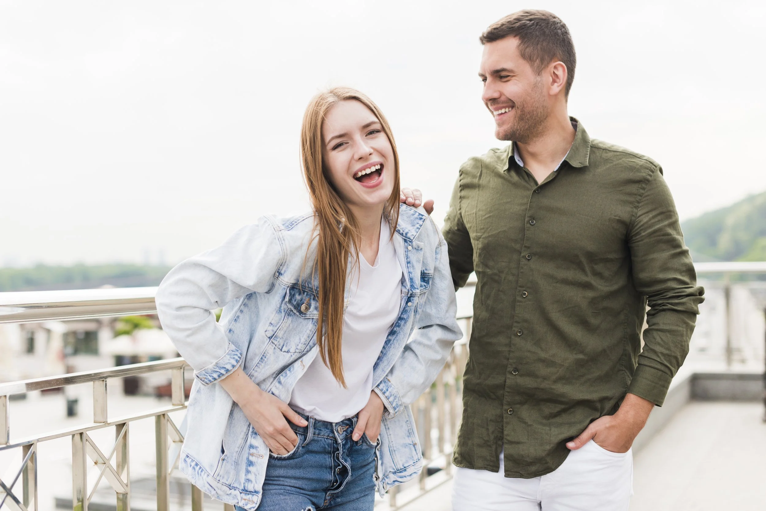 A young woman with long red hair and a man with short dark hair smiling, standing outdoors on a balcony with a cityscape in the background. The woman is wearing a denim jacket and white t-shirt, with her hand on her hip, while the man is wearing a green button-up shirt and white pants, with his arm around the woman.