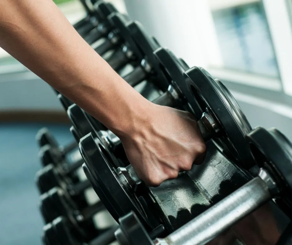 Close-up of a person’s hand gripping a black dumbbell on a rack in a gym.