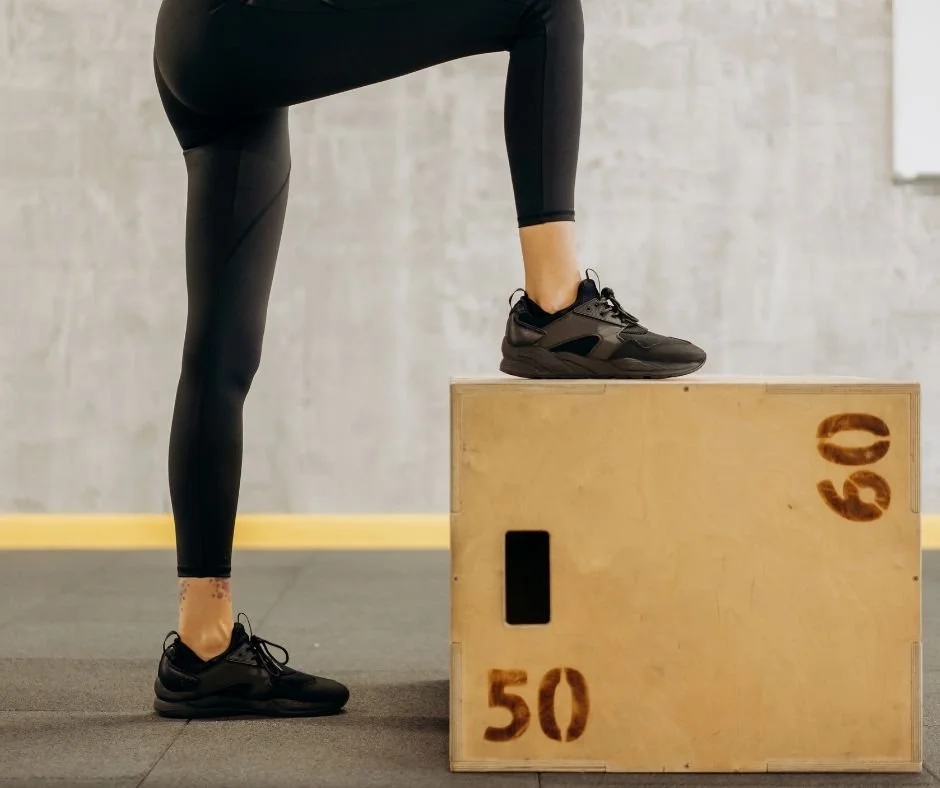 Person in black leggings and athletic shoes standing with one foot on a wooden plyometric box marked '50' and '60', in a gym setting.