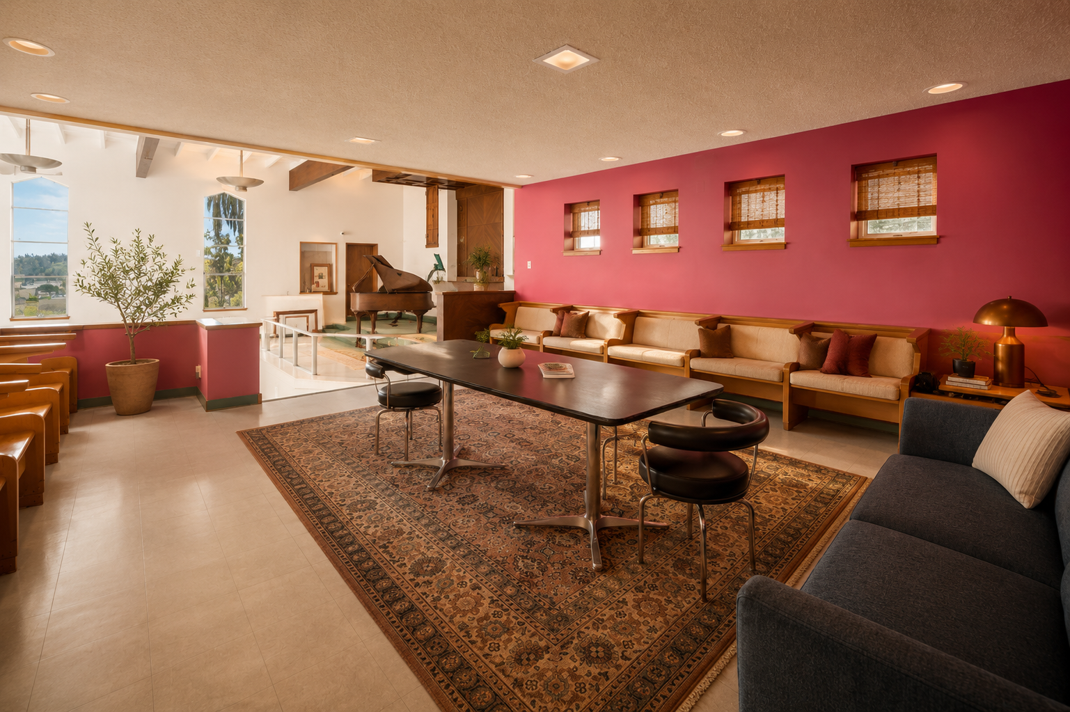 A cozy reading and music lounge with a pink accent wall, beige seating, a dark wood table, and a grand piano near white and red walls, decorated with plants and framed pictures.