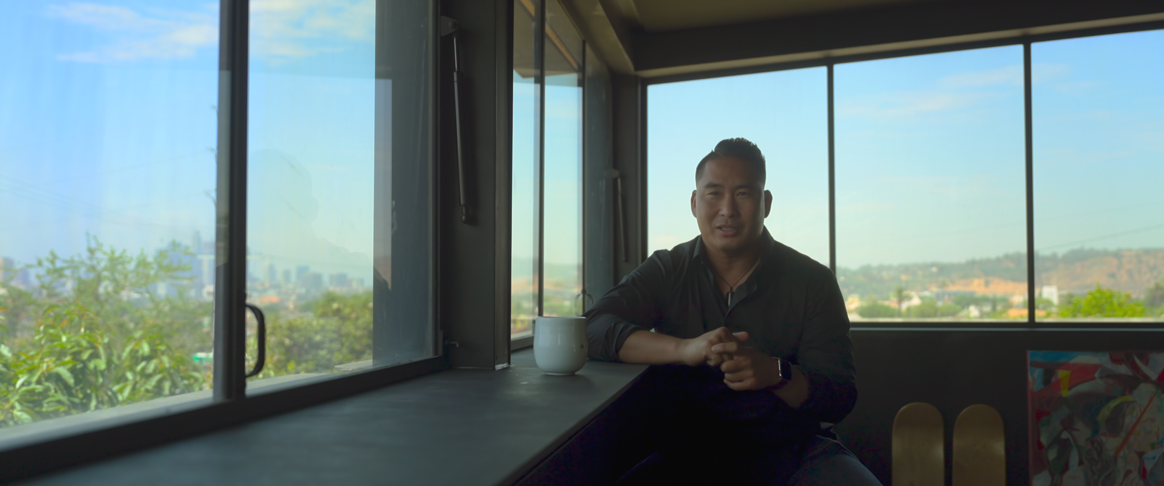 A man sitting at a counter inside a room with large windows overlooking a cityscape and distant hills, holding his hands together near a white mug.