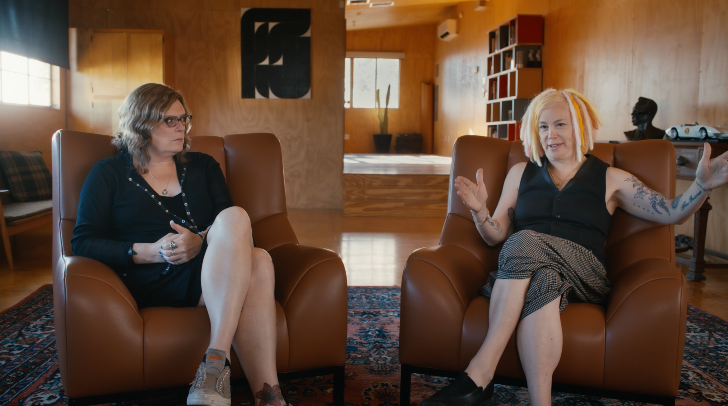 Two women sitting on brown leather chairs in a room with wooden walls, engaged in conversation. One woman has blond dreadlocks and tattoos, the other has shoulder-length brown hair and glasses.