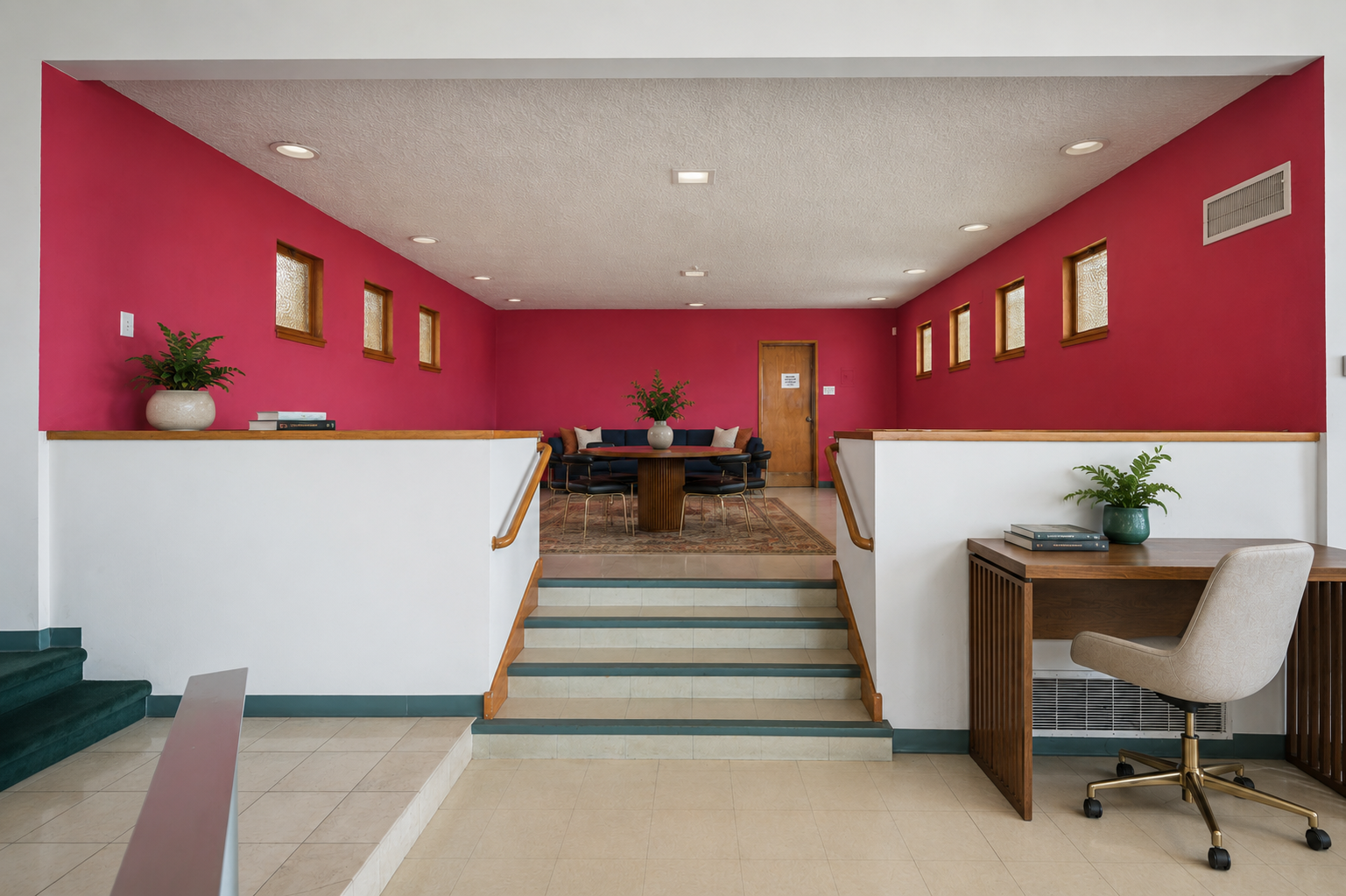 Interior of a restaurant or cafe with a pink accent wall, small windows, wooden accents, potted plants, a round table with black seating, and a office chair at a wooden desk.