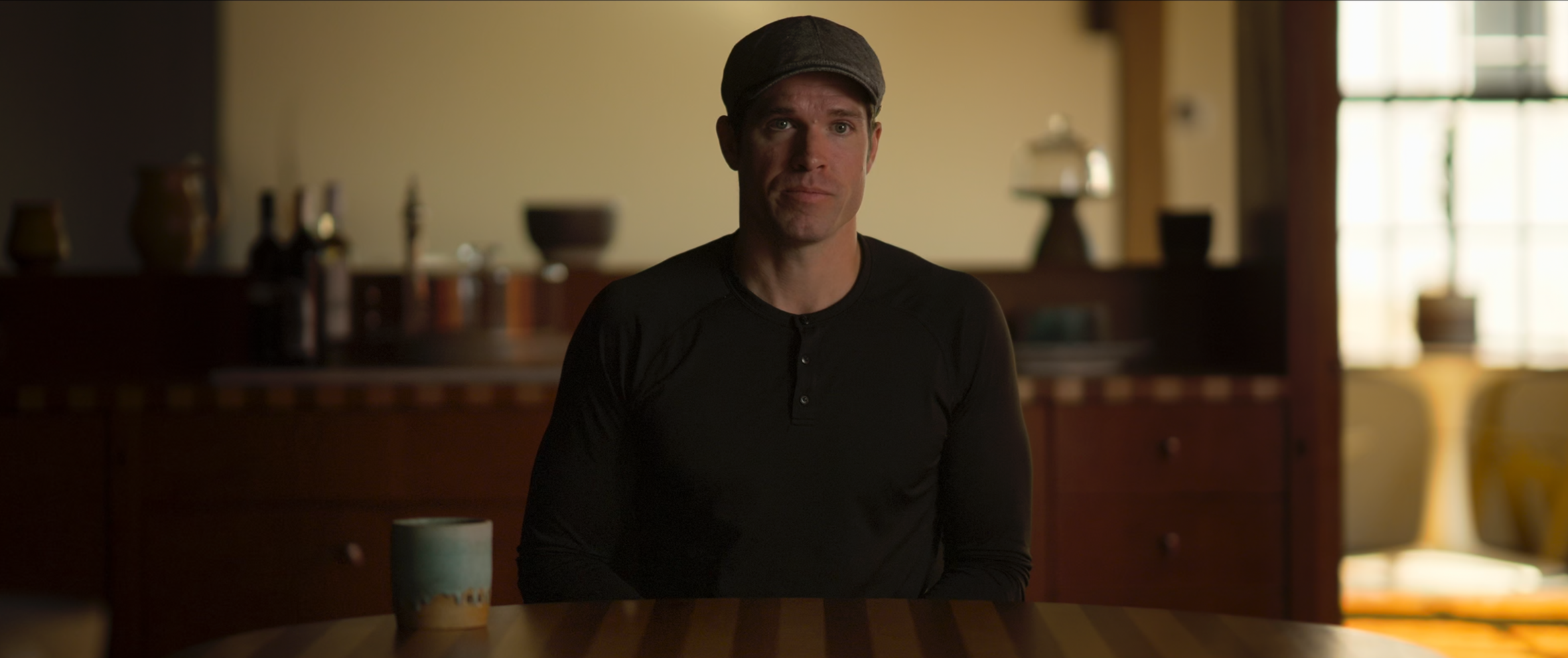Jackson Spidell sitting at a dining table in a dimly lit room, wearing a black shirt and a gray cap. Wick is Pain. Narrator Studios