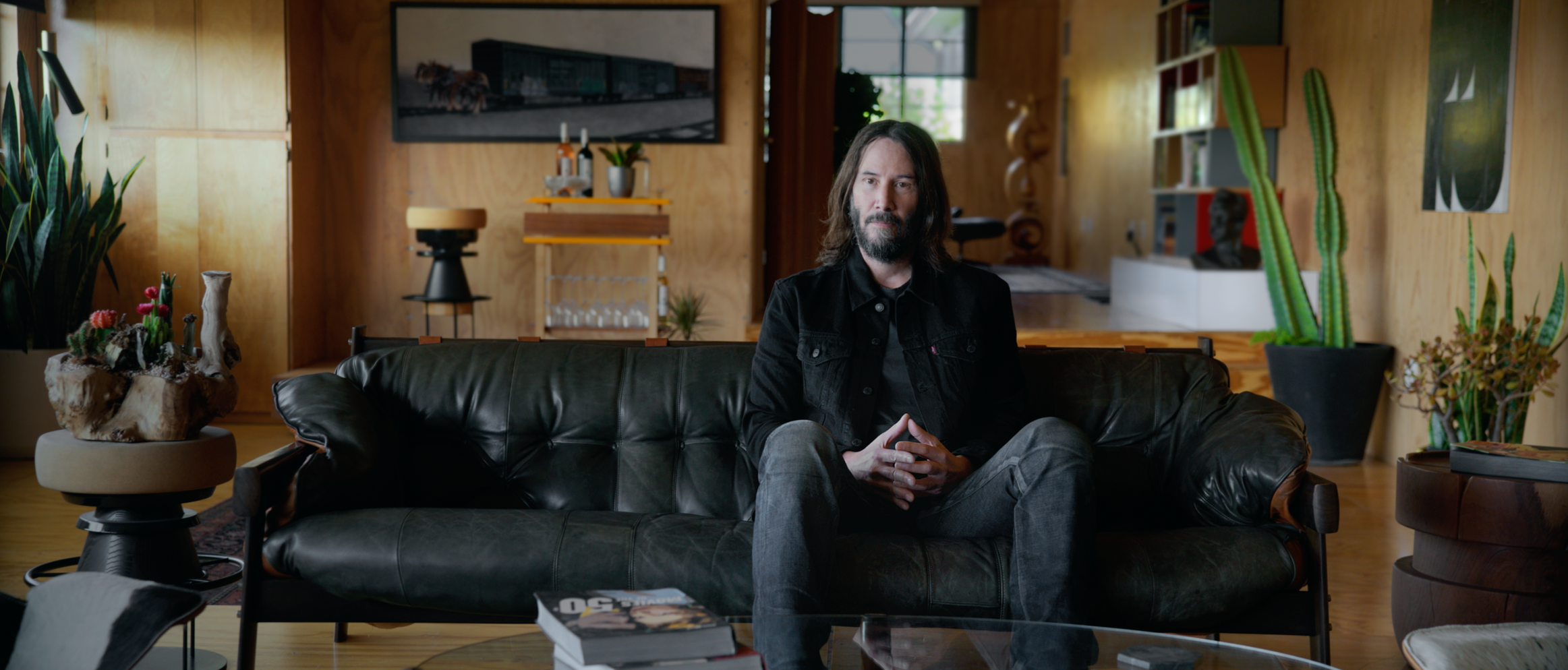 Keanu Reeves sitting on a black leather sofa in a modern living room, surrounded by plants and wooden decor. Narrator Studios.
