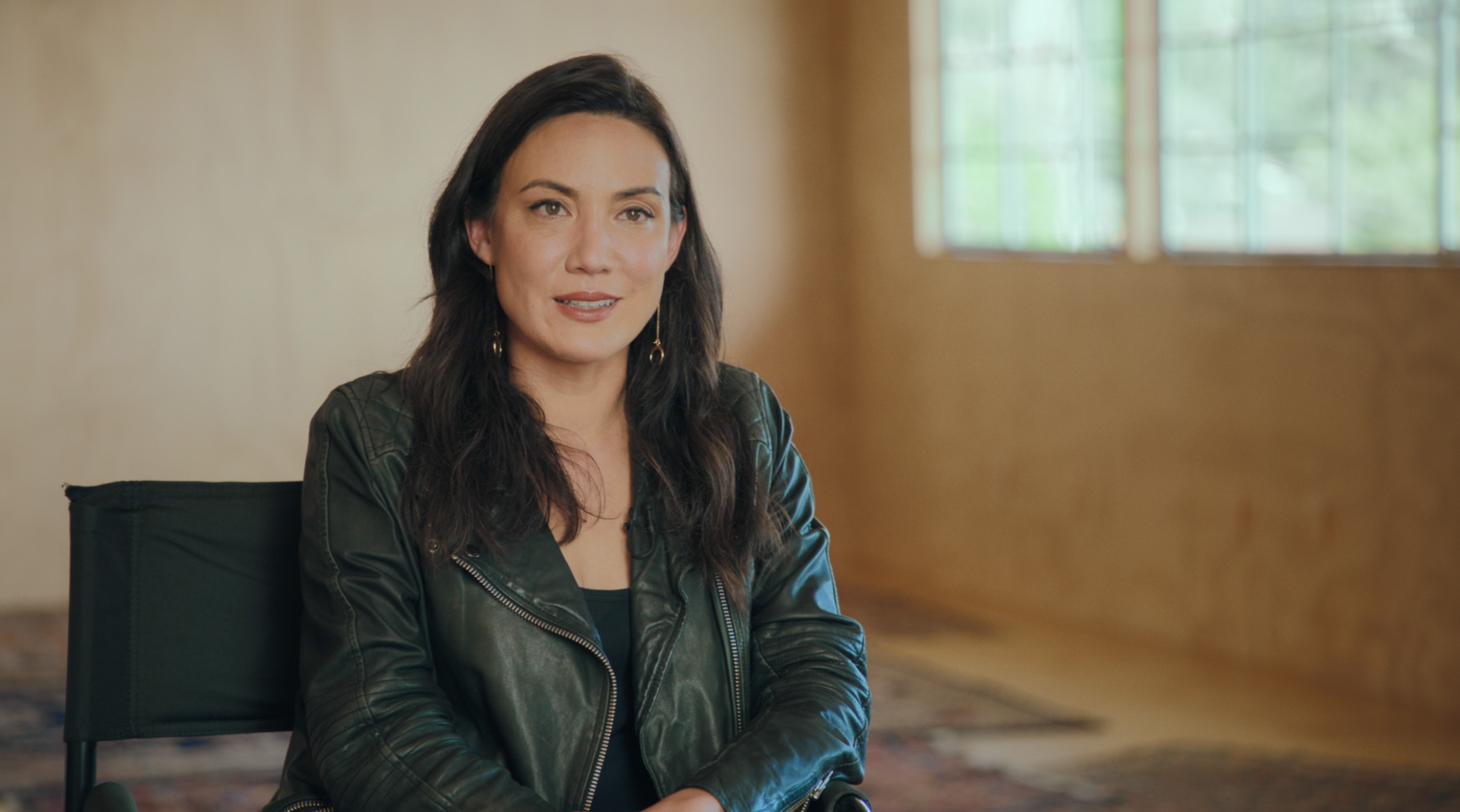 A woman with long dark hair, wearing a black leather jacket, sitting on a black chair in a room with beige walls and a large window with sunlight streaming in.