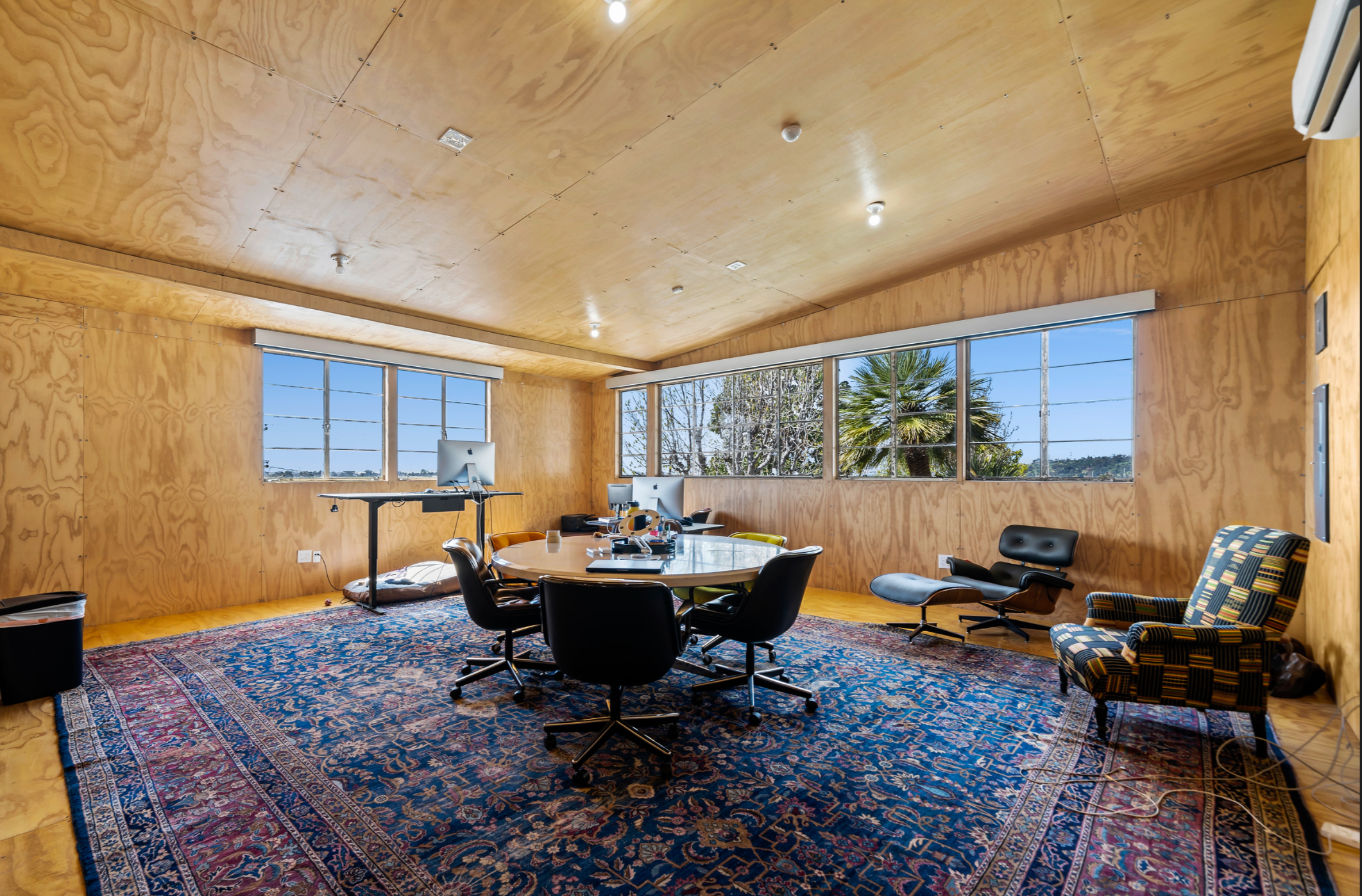 Office space with wooden walls and ceiling, large windows showing a view of trees and a blue sky, a patterned rug on the wooden floor, a round table with multiple chairs, an ergonomic lounge chair, a colorful armchair, and computer equipment.
