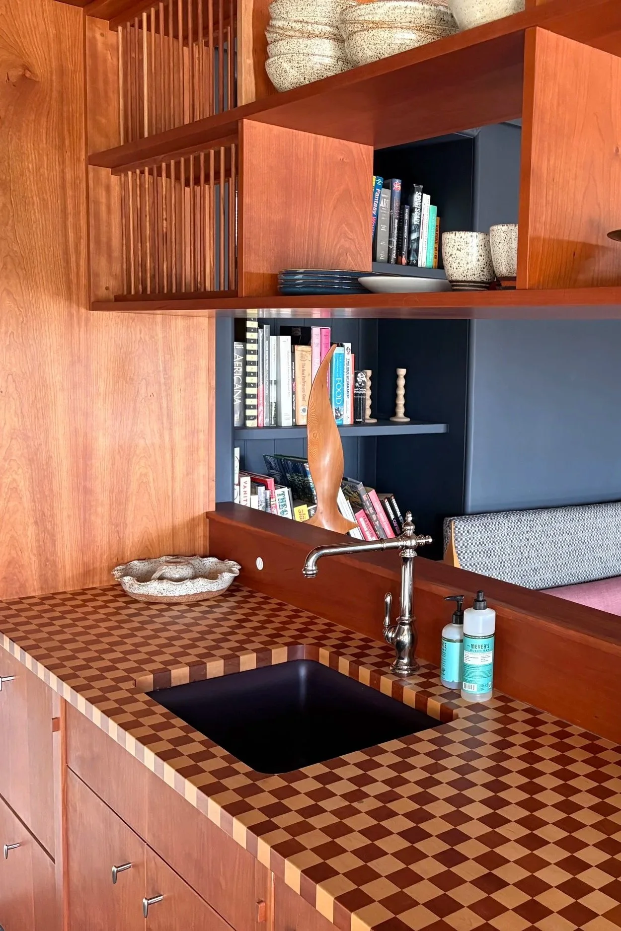 Kitchen with wooden cabinetry, a countertop with a checkered pattern made of small wood squares, a black sink, a silver faucet, and soap bottles. Behind the counter are open shelves with books, dishes, and decorative items, including ceramic cups and candle holders.