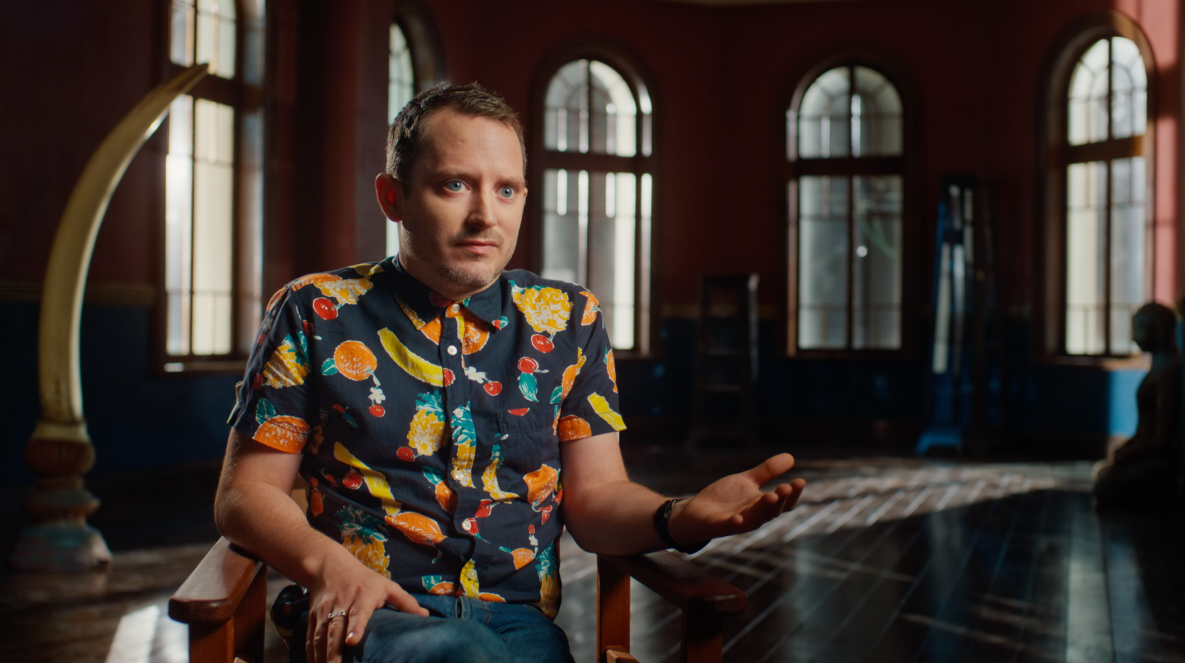 Elijah Wood wearing a colorful short-sleeve button-up shirt with a fruit pattern, is sitting on a wooden chair in a room with large arched windows. He appears to be engaged in a conversation or in thought, with his hands gesturing slightly.