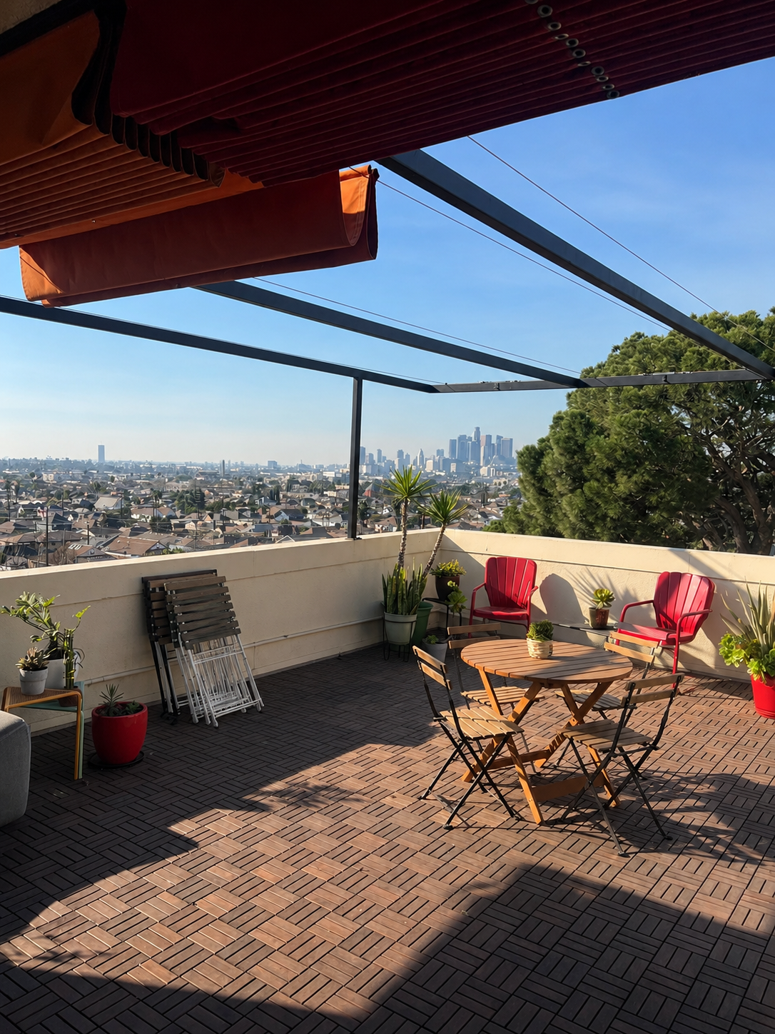 Rooftop patio with wooden furniture, potted plants, and an expansive cityscape view of Los Angeles skyline in the distance.