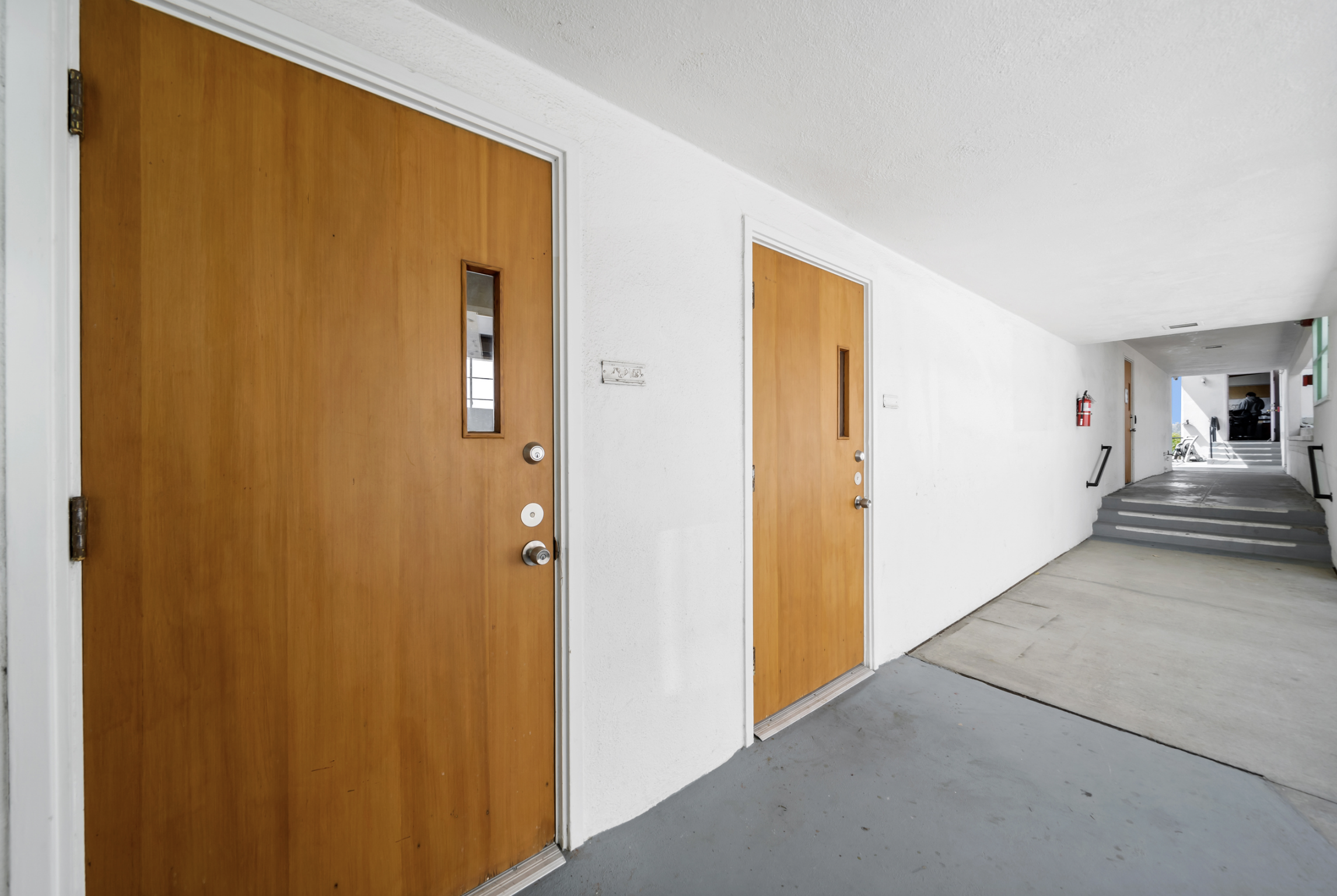 Hallway with wooden doors and white walls, fire extinguisher on wall, stairs leading up and a person sitting in background.