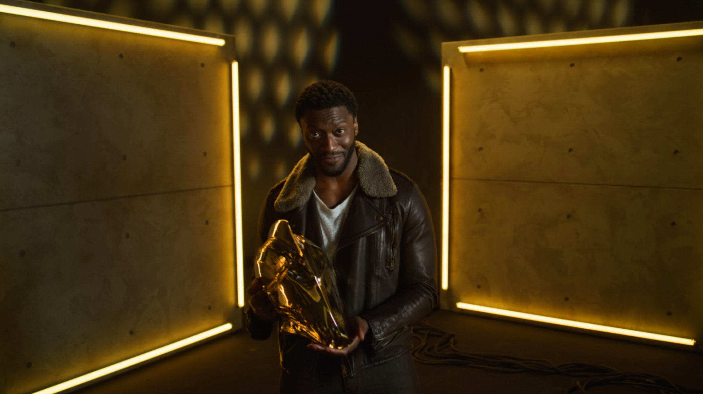 A man wearing a leather jacket with a fur collar, stands in a dimly lit corridor with yellow neon lights and holds a shiny gold object.