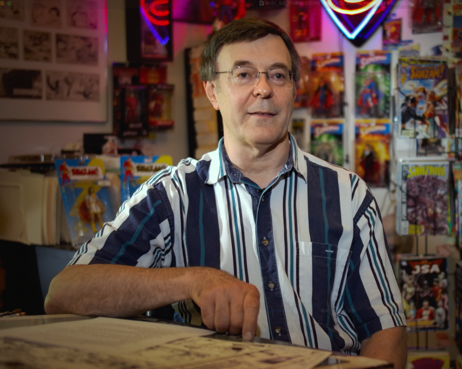 A man with glasses wearing a striped shirt sitting at a table in a comic book store, with comic book racks and posters in the background.