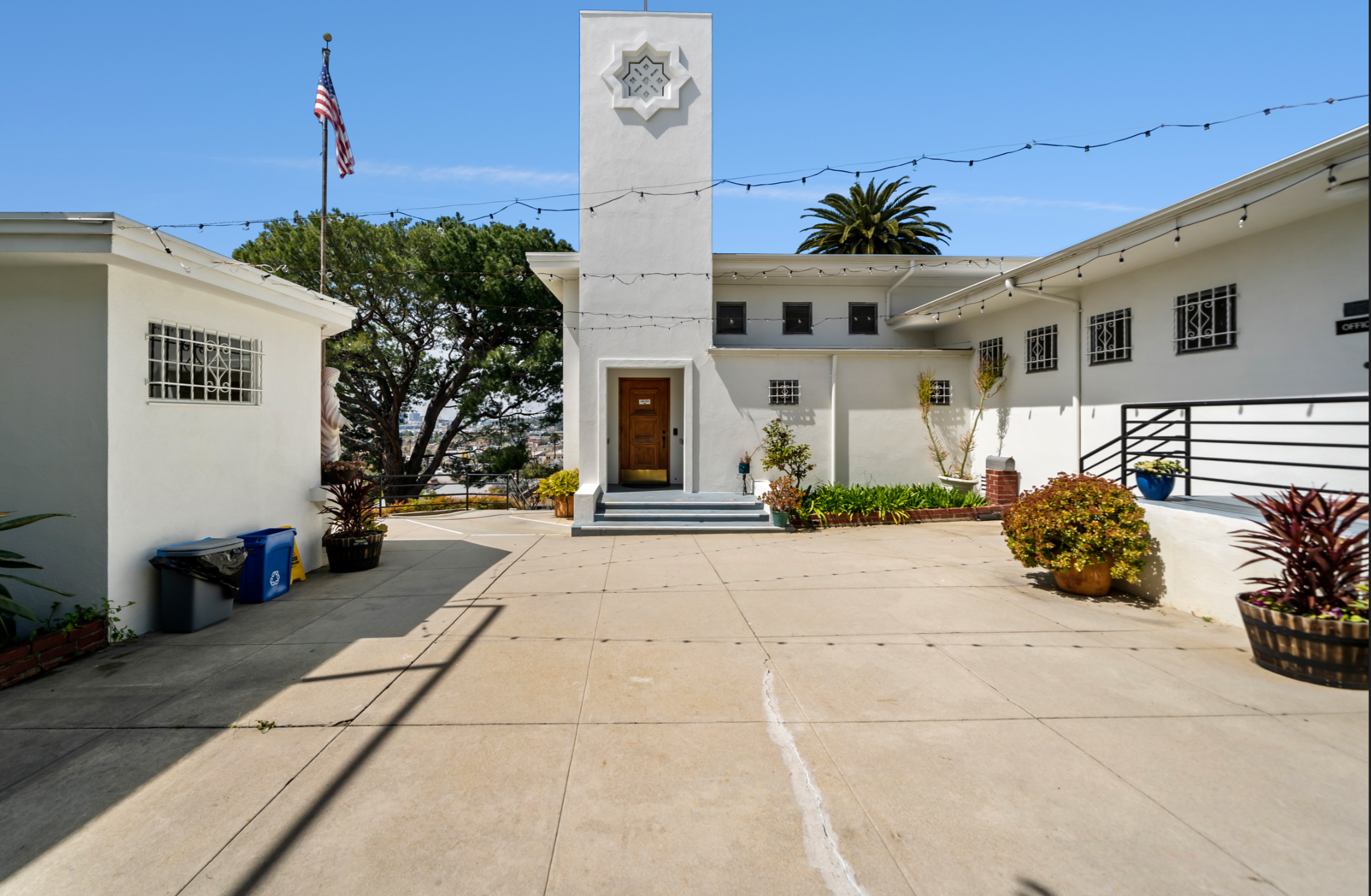White building with a tower and a star-shaped window, outdoor patio with potted plants, string lights, American flag, and trees in the background.