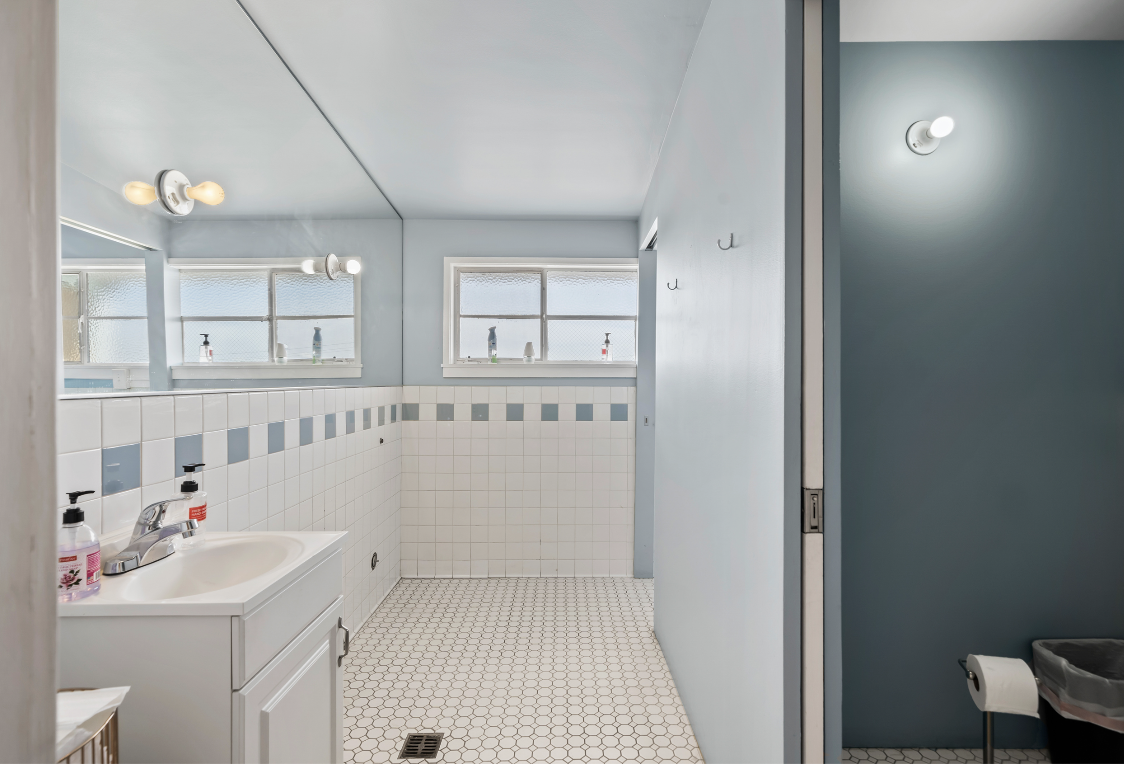 A small bathroom with white and blue tiled walls, a white vanity with a sink, soap dispensers, and bottles, two windows with frosted glass, and overhead lighting. There is a doorway to a separate area with a trash can visible.