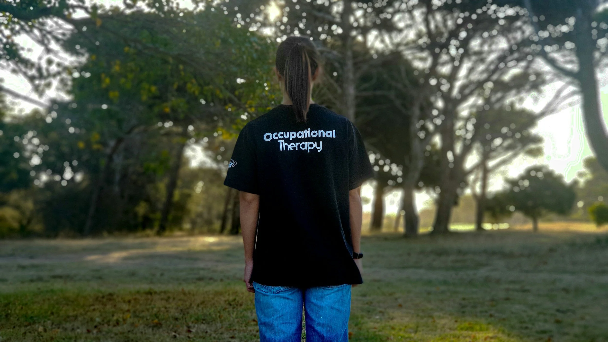 A person with dark hair tied back, wearing a black T-shirt with white text that says 'Occupational Therapy,' stands outdoors in a park surrounded by trees and sunlight filtering through the branches.