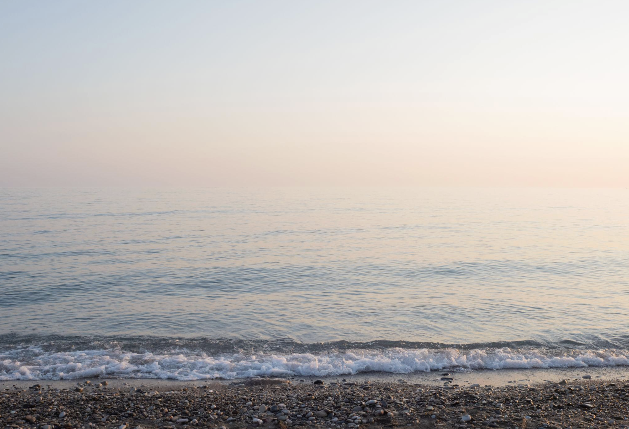 A calm ocean with gentle waves lapping against a pebble-covered shoreline, under a clear sky with soft pastel hues.