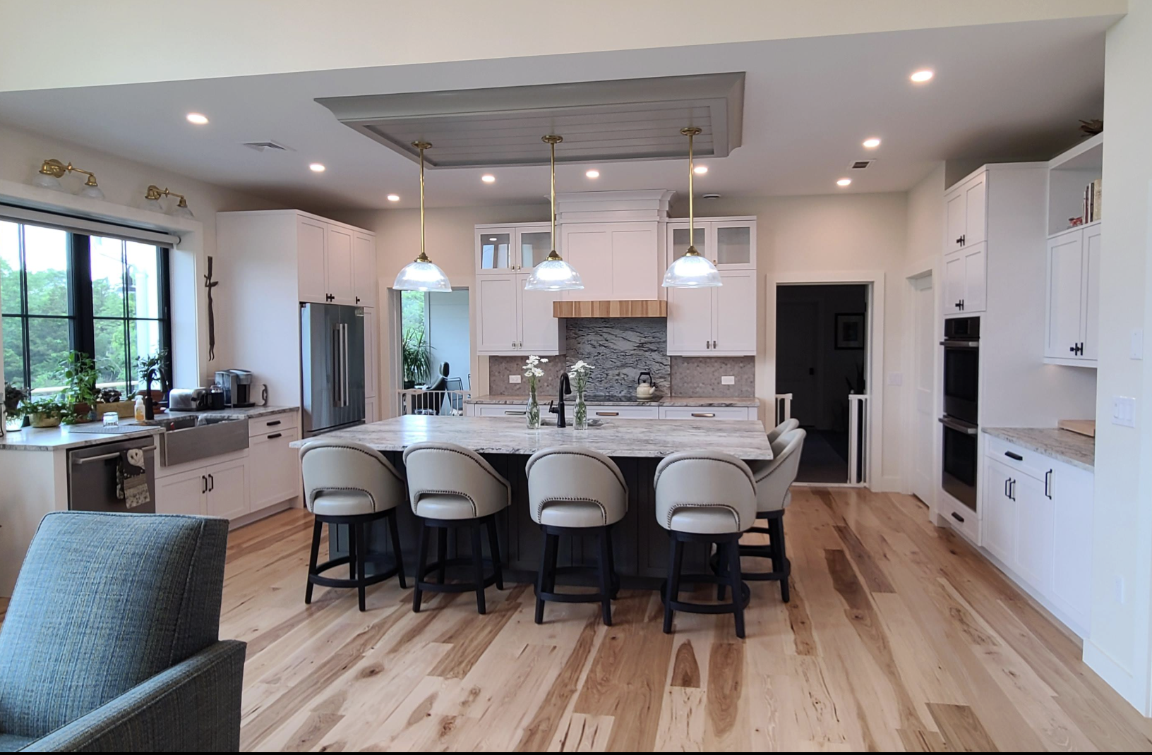 Modern kitchen with white cabinets, marble countertops, and a large island with six cream-colored chairs. There are three pendant lights above the island, a window with plants, and hardwood floors.