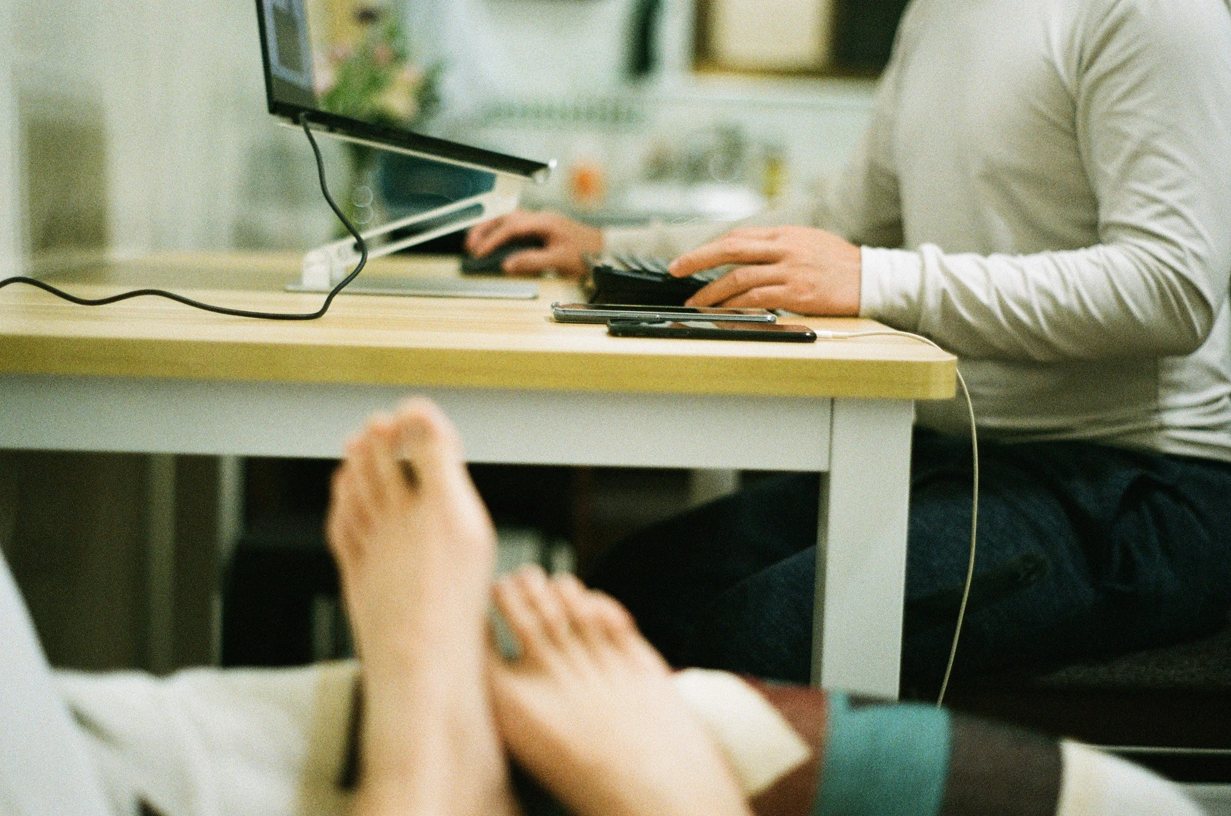 A person in a white long-sleeve shirt working on a computer at a desk with two smartphones and a smartwatch on the desk, with another person's hand visible in the foreground.