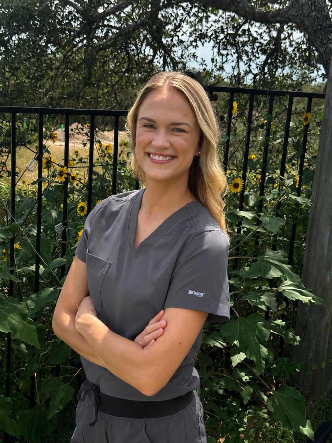 A woman with blonde hair smiling with arms crossed, wearing gray medical scrubs, standing outdoors in front of a black fence with yellow sunflowers and greenery.