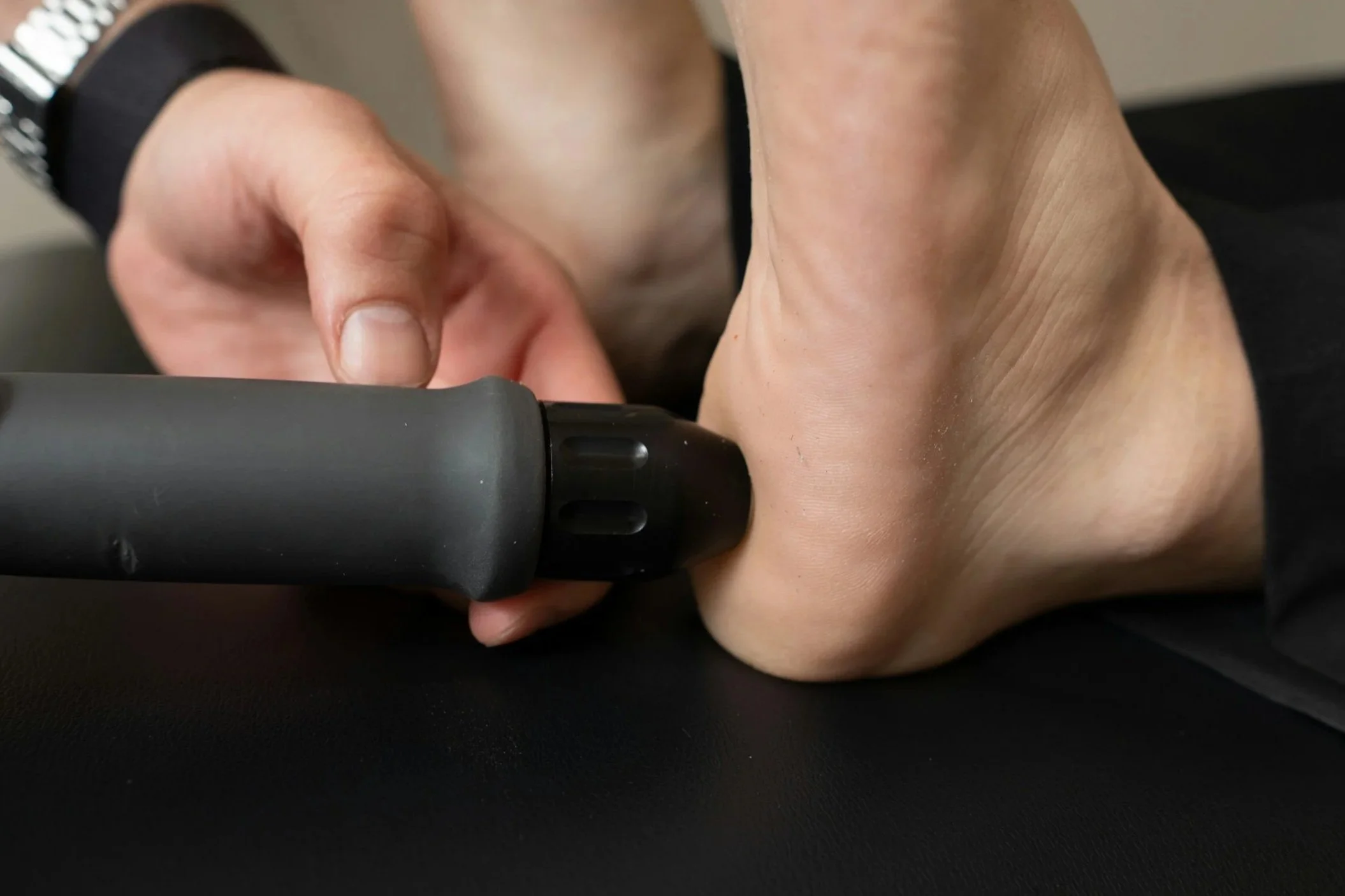Close-up of a person holding a black flashlight or tool, pointing it downward, with focus on their hand and wrist. The person is wearing a dark sleeve.