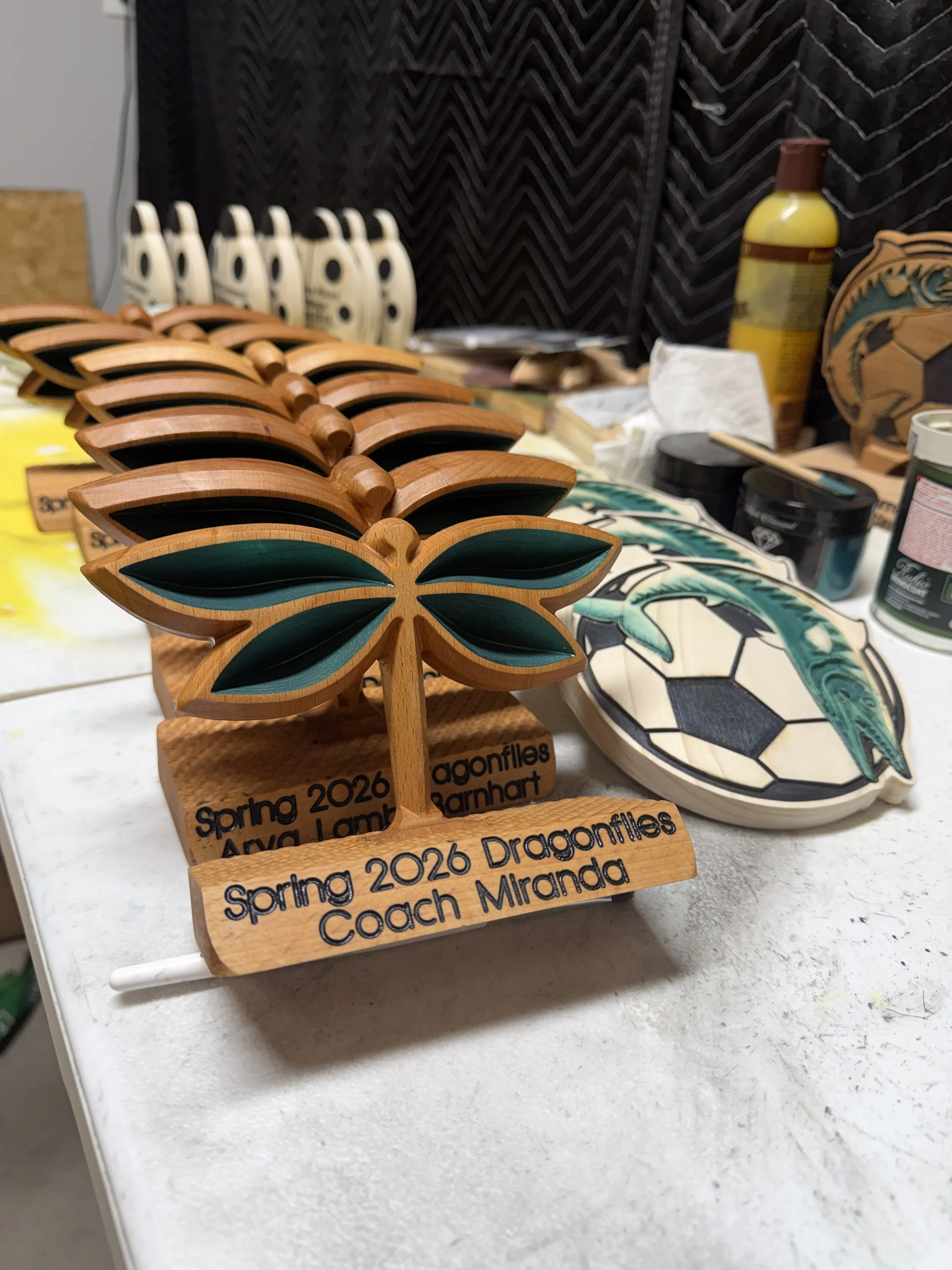 Wooden awards shaped like dragonflies, engraved with 'Spring 2026 Dragonflies Coach Miranda,' placed on a table among various art and craft supplies, with a dark fabric backdrop.