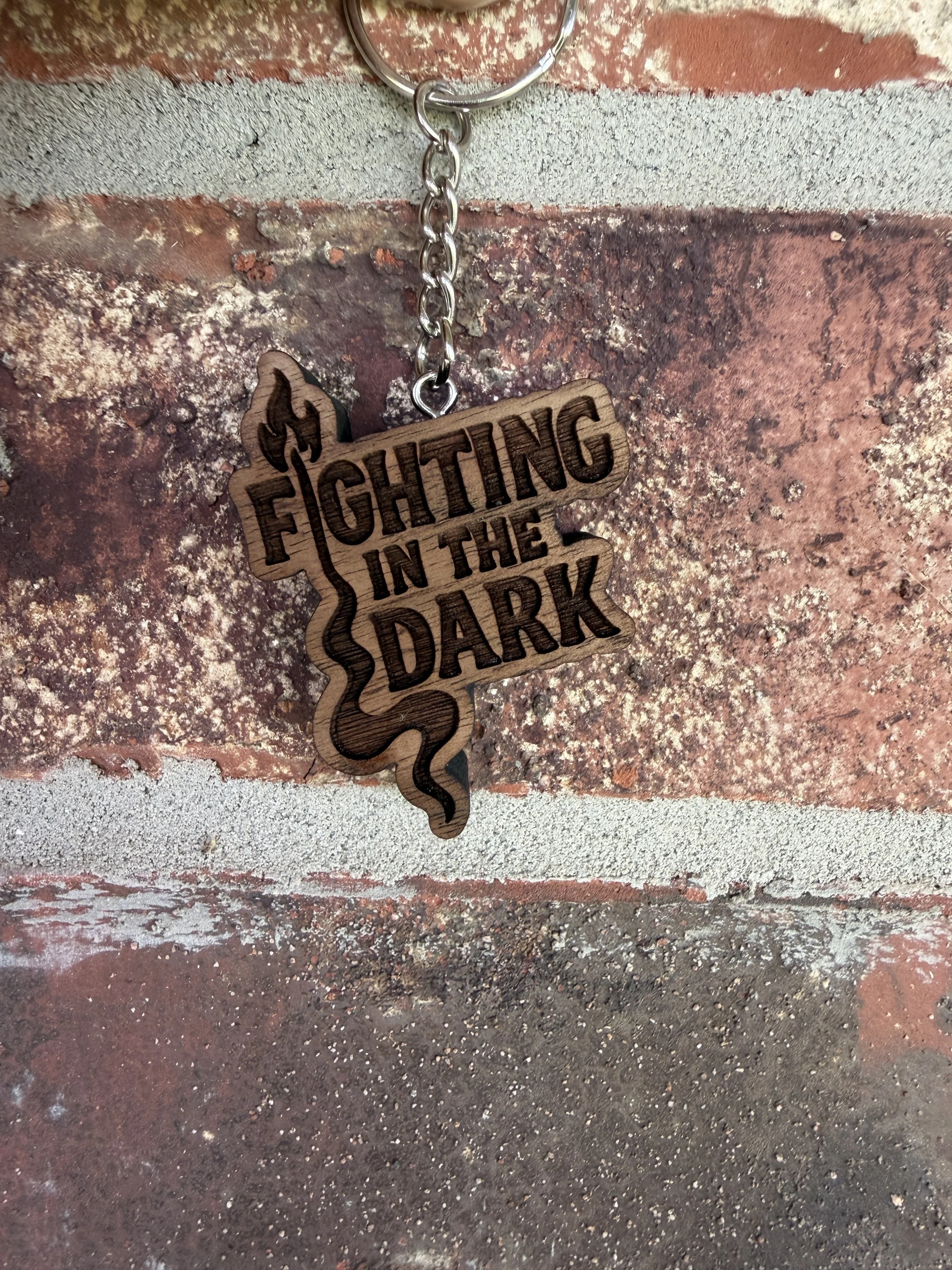 Wooden keychain with a cutout of the state of Texas and the phrase 'Fighting in the Dark' engraved on it, hanging from a metal keyring attached to a brick wall.