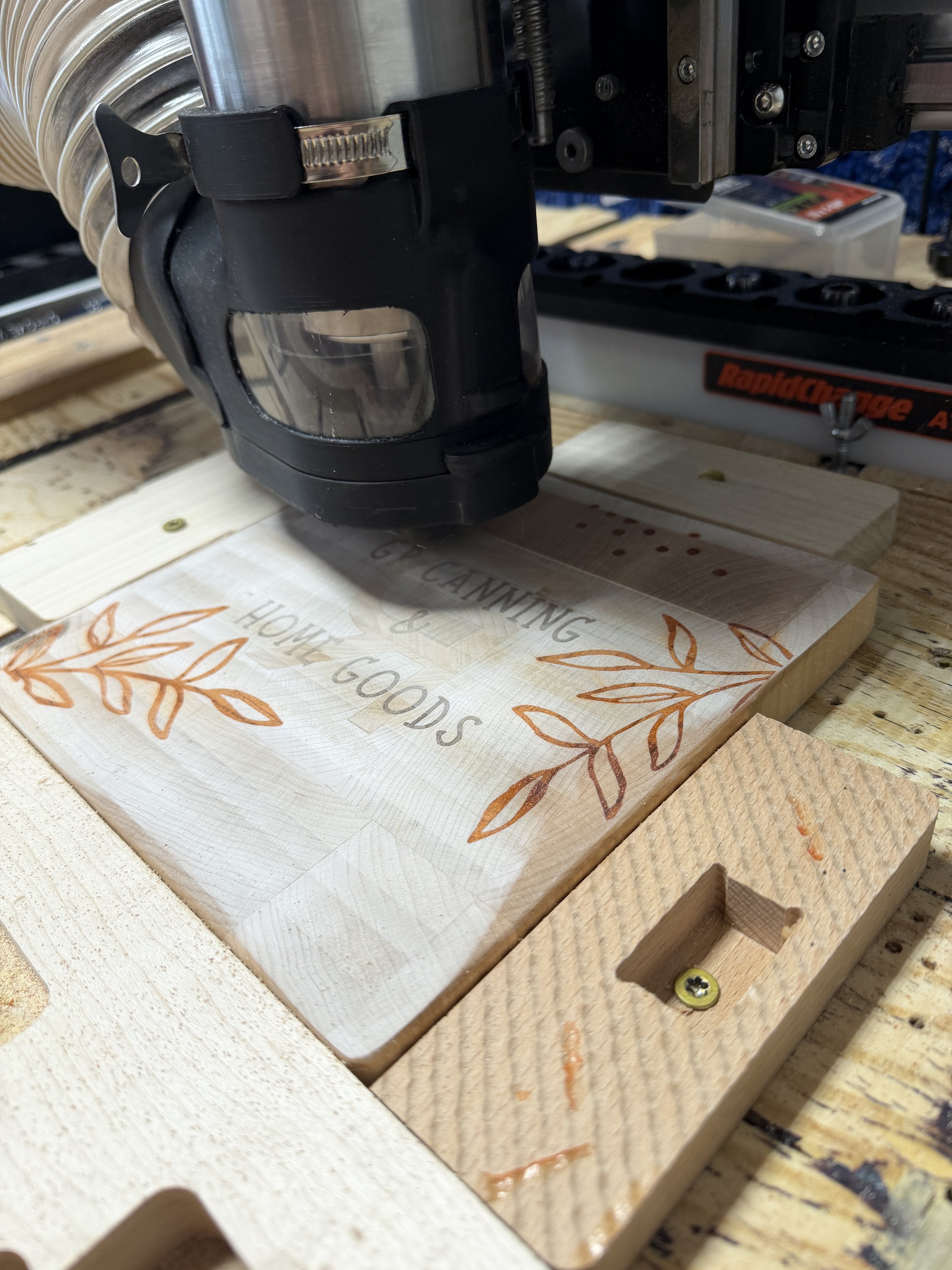 A woodworking scene with a piece of wood that has a decorative leaf pattern and the words 'HOME COOKS' carved into it, being cut or engraved by a power tool.