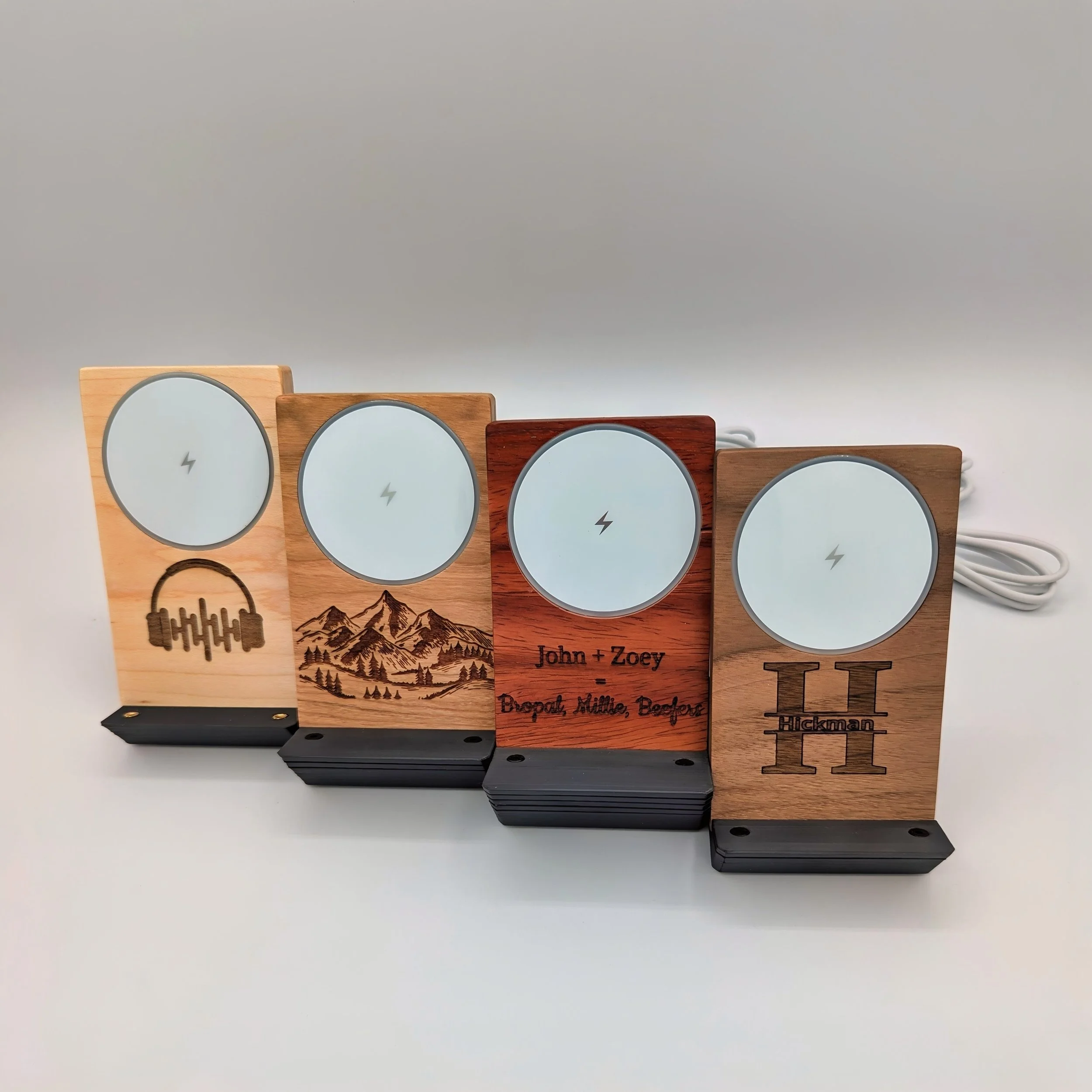 Four wooden wireless chargers with engraved designs and text, placed on a white surface against a plain gray background.