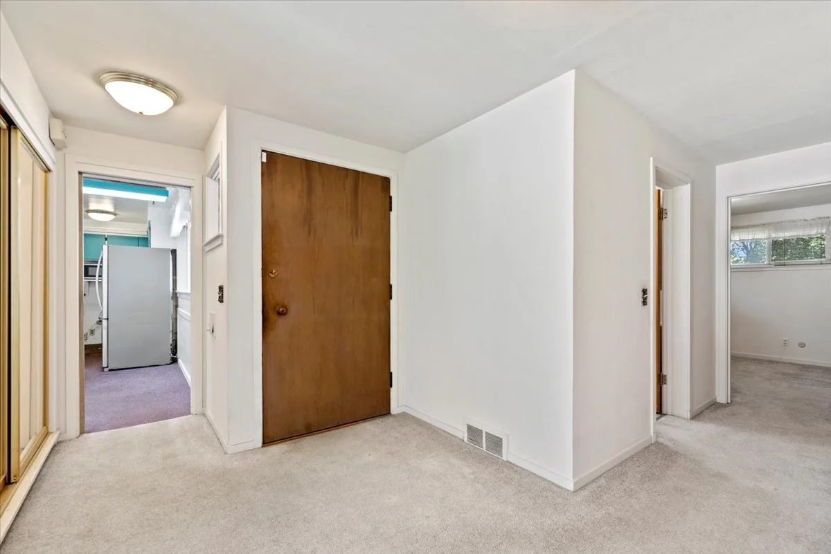 Interior view of a home showing a hallway with a wooden front door, closet with mirrored sliding doors, and doorways leading to other rooms, with beige carpet and white walls.