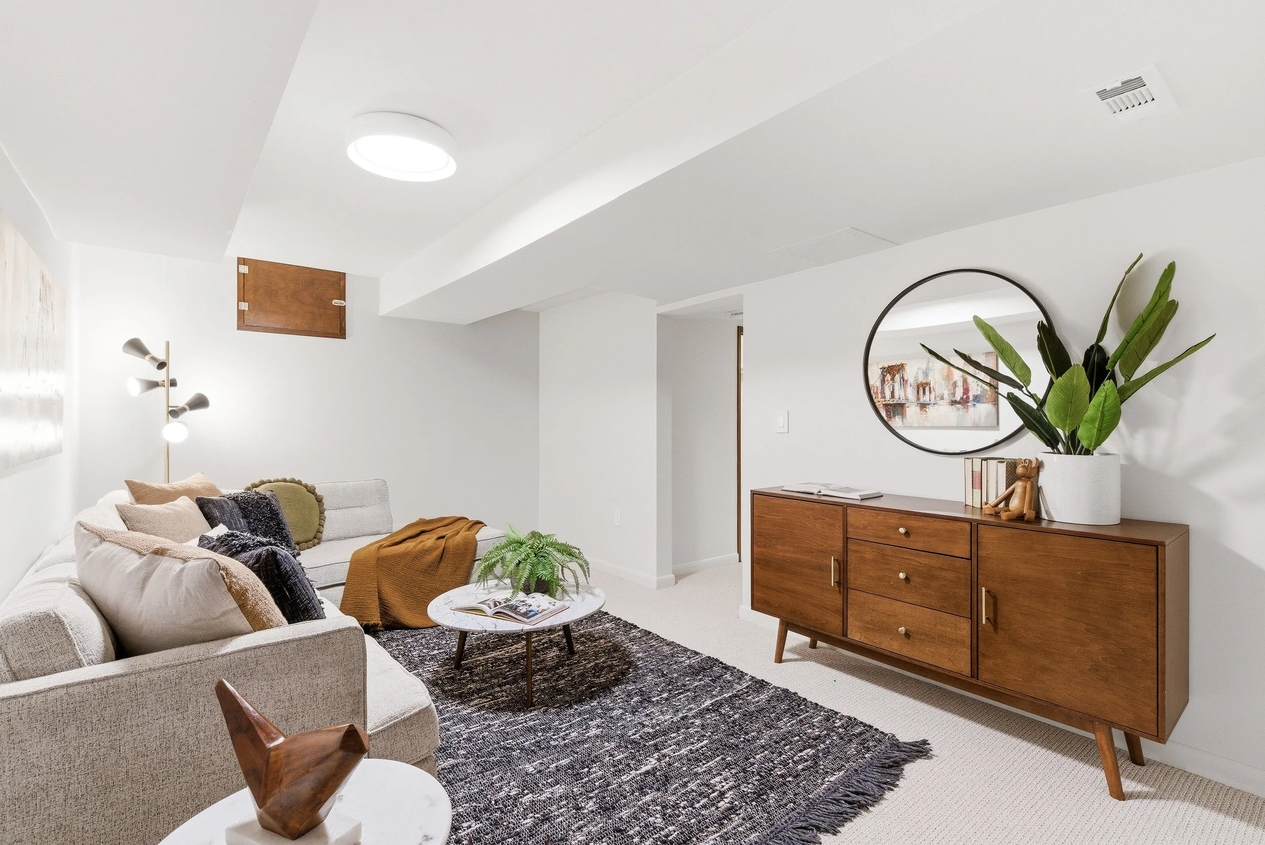 Living room with a beige sofa, throw pillows, a small round coffee table with a plant and open magazine, a black and white patterned rug, a wooden sideboard with a plant and artwork, white walls, a large round mirror, a floor lamp, and ceiling lights