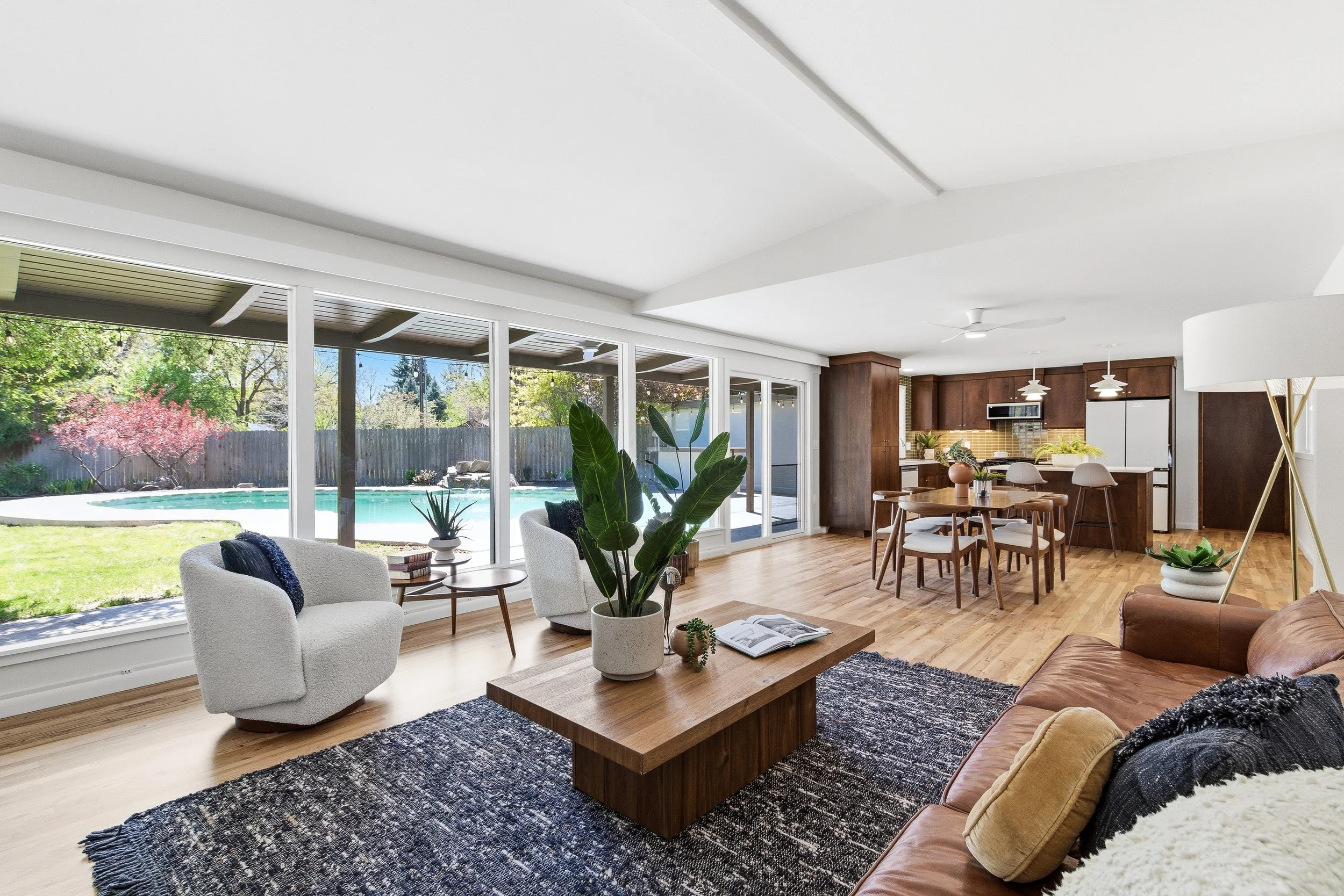 Spacious open-plan living room and dining area with large windows and sliding glass doors overlooking a backyard with a swimming pool, trees, and a wooden fence. The room features white walls, wooden flooring, a tan leather sofa, white armchairs, a w