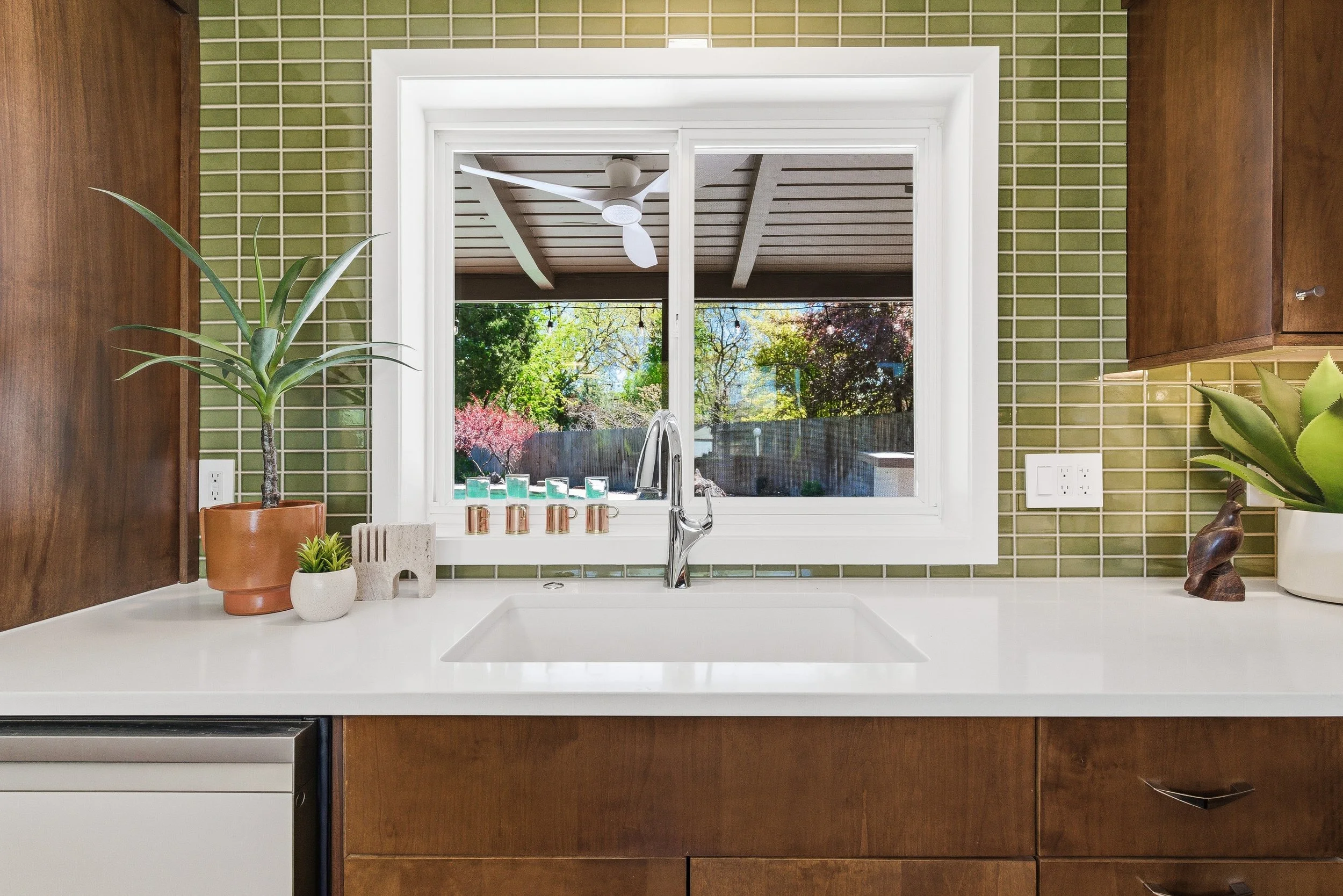 View from inside a kitchen, looking out through a window at a backyard with trees and a fence, with a ceiling fan visible outside.