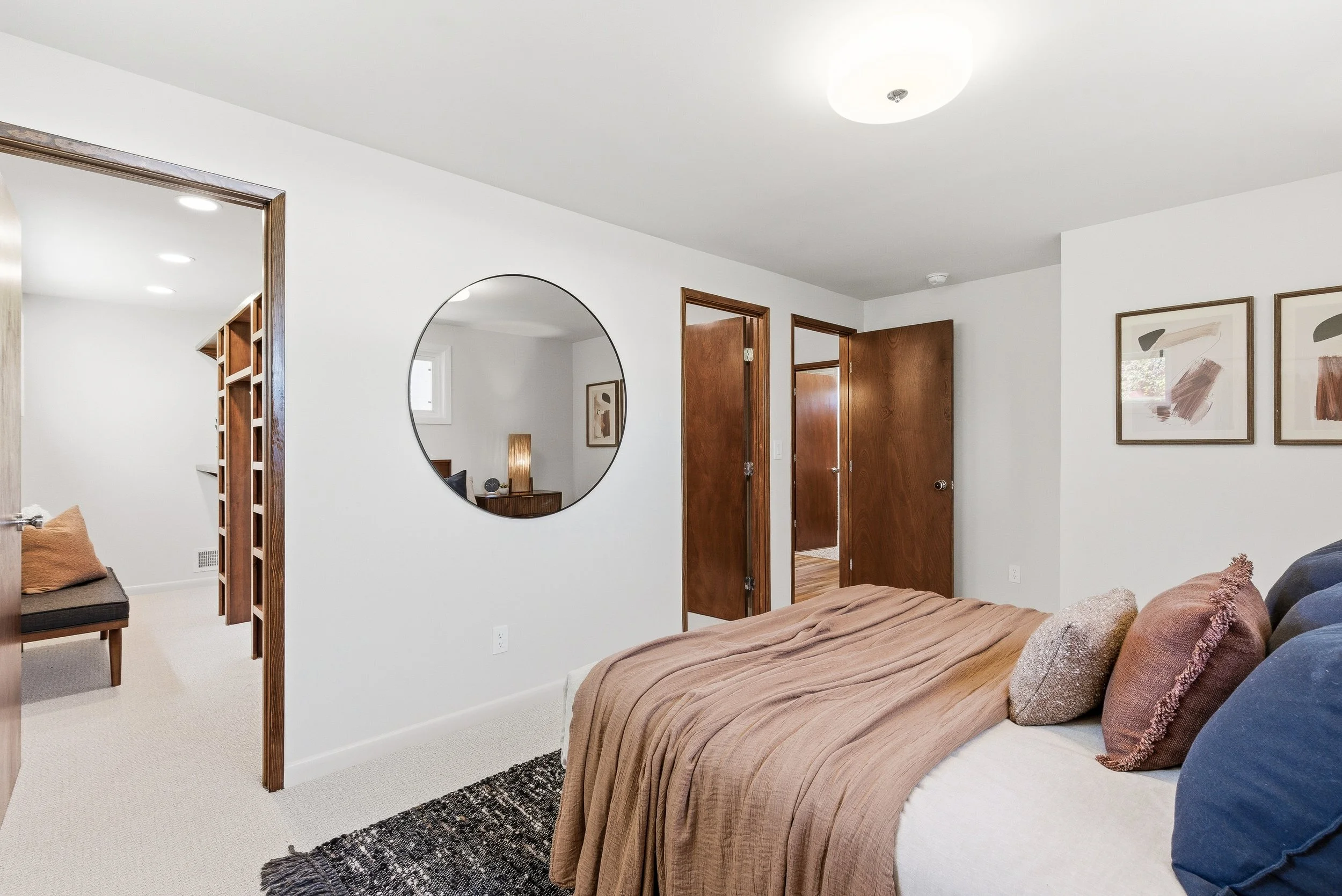 A cozy bedroom with a bed covered in a brown blanket, decorative pillows, and a textured black and white rug. A large round mirror hangs on the white wall, reflecting a small window and a nightstand with a lamp. There are wooden doors leading to othe