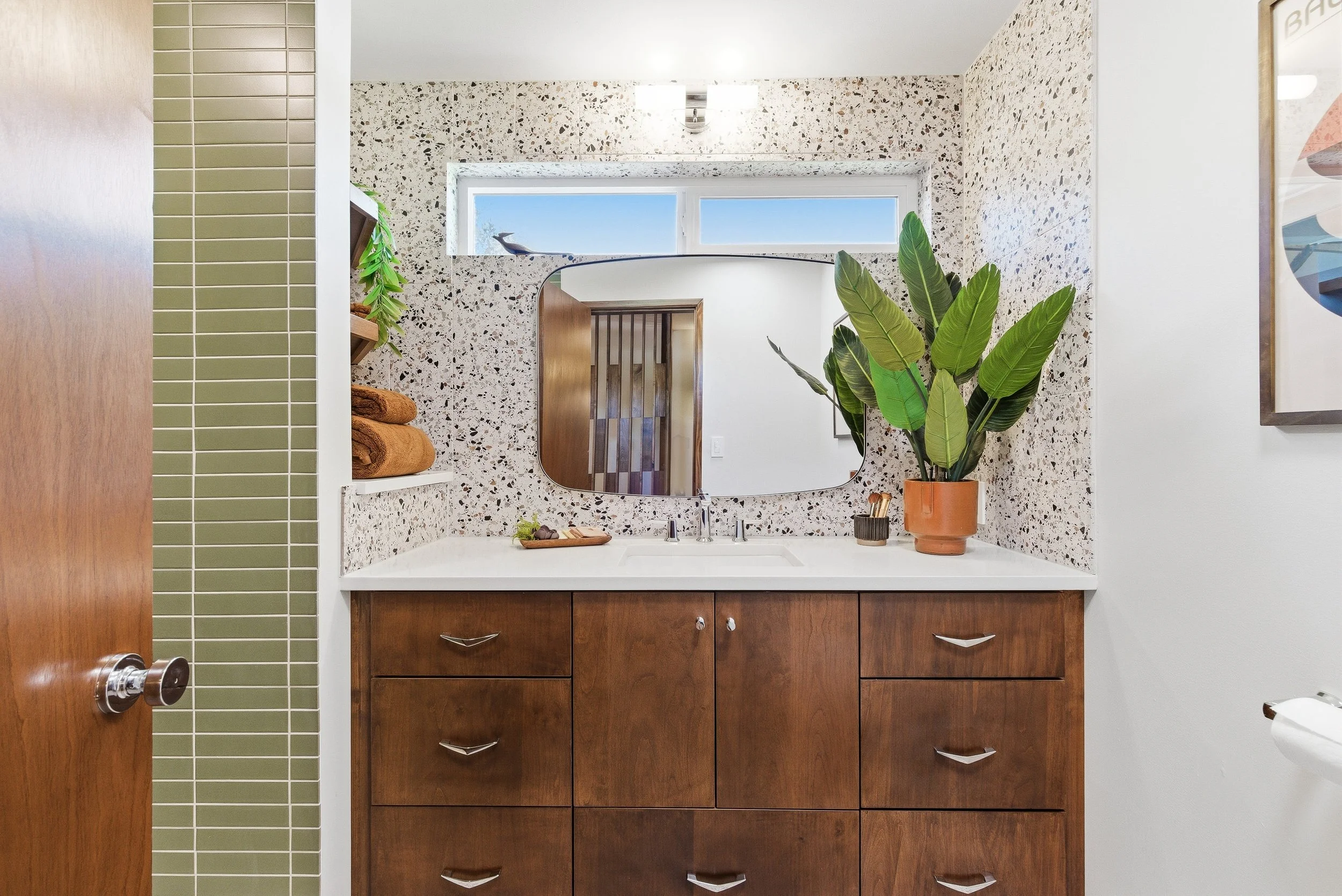 Bathroom sink area with a terrazzo wall, wooden cabinetry, a large mirror, a potted plant, and a window with a bird outside.