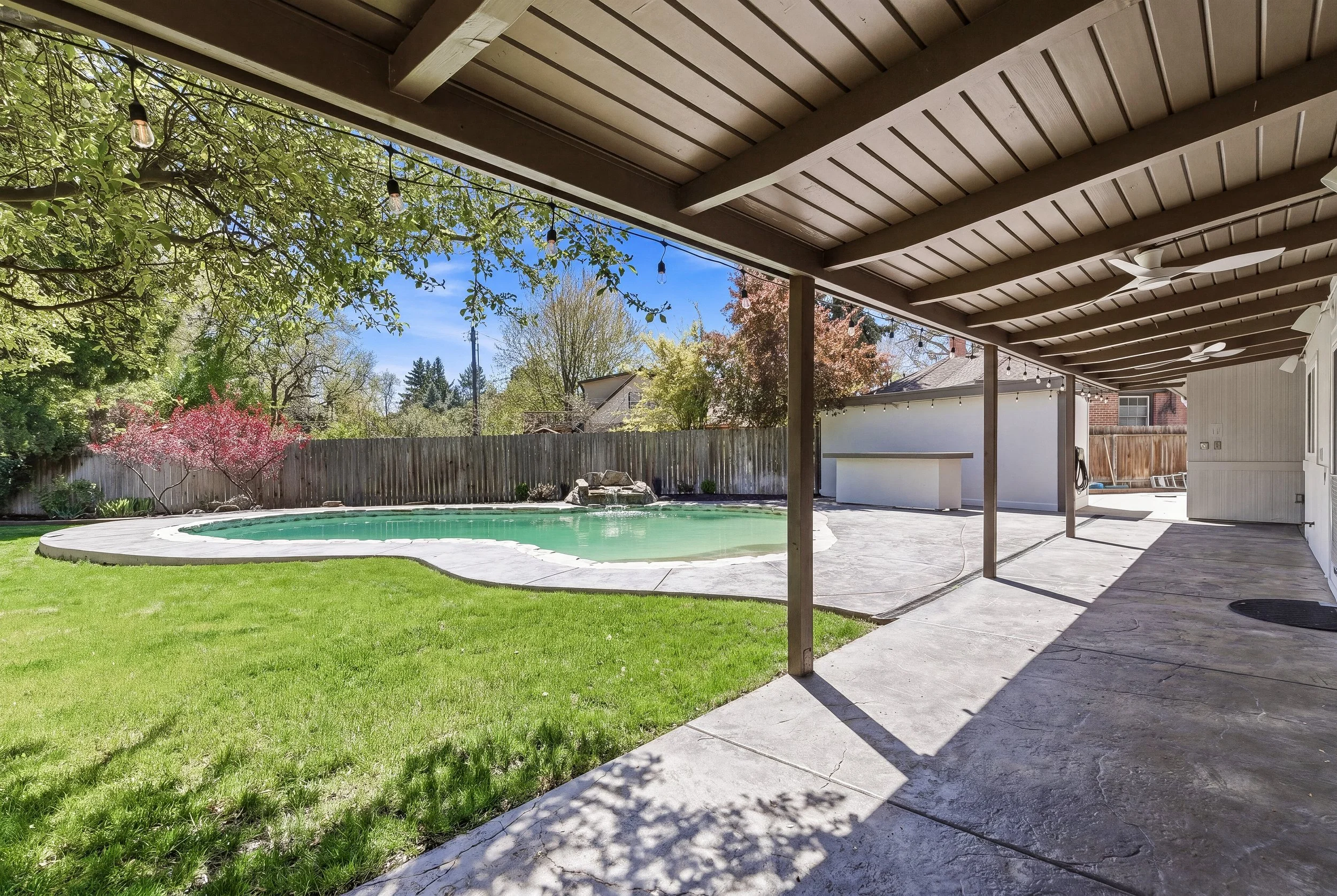A backyard scene with a swimming pool, green grass, trees, and a wooden fence. There is a covered patio area with string lights hanging from the ceiling.