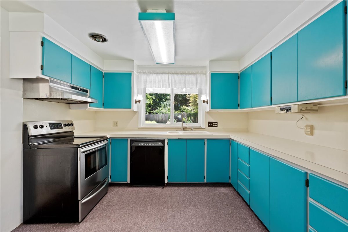 Kitchen with white countertops, bright turquoise cabinets, stainless steel oven and stove, black dishwasher, window with white curtains, and a view of greenery outside.