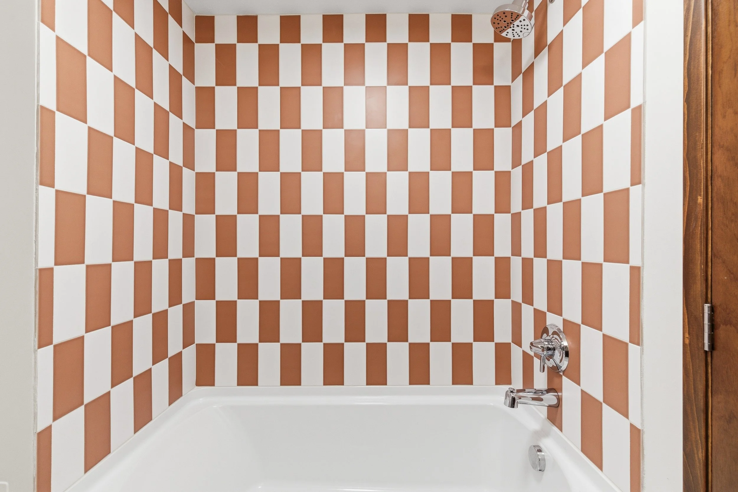 A shower with brown and white checkered tiles on the walls, a round chrome showerhead in the top right corner, and a chrome faucet and handle at the bottom right, with a white bathtub at the bottom.