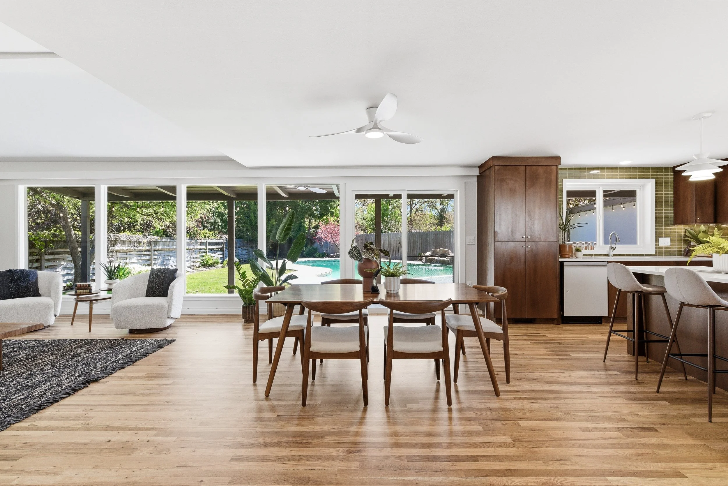 Open-concept living room and kitchen with large windows, wooden dining table, white chairs, and plants, overlooking backyard and pool.
