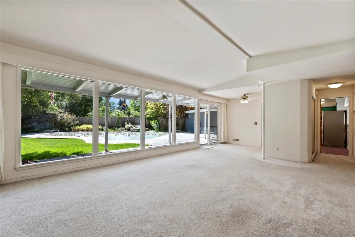 Empty living room with large glass windows overlooking a backyard with a pool and greenery.
