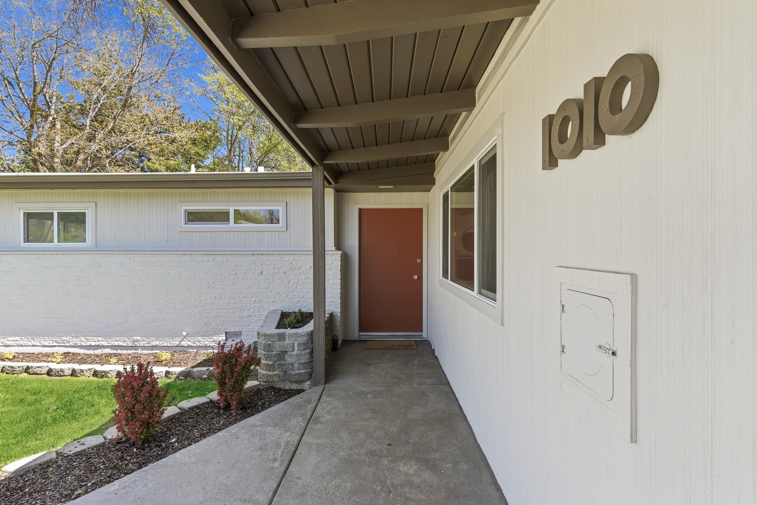 A residential house entrance with a red door, concrete walkway, decorative brick planter with plants, white exterior walls with horizontal siding, a window with a screen, and the letter 'P' mounted on the wall. Part of the landscaped yard with green 