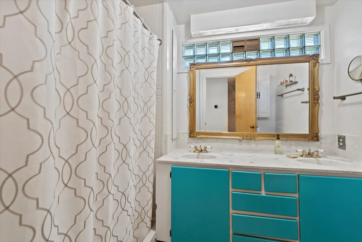 Bathroom with double sink, marble countertop, large ornate gold-framed mirror, blue cabinets, shower with patterned curtain, small window with glass blocks, and white walls.