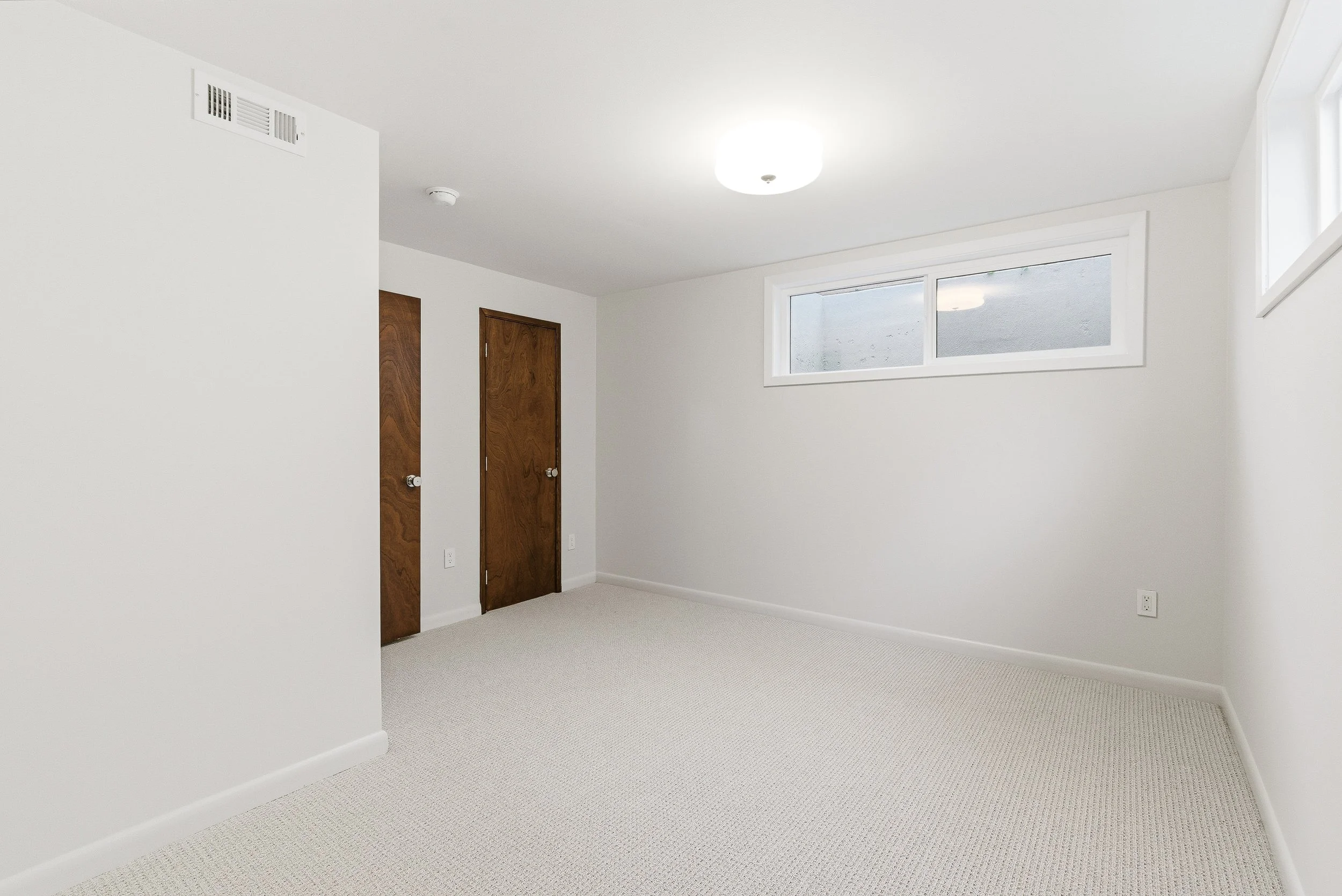 Empty white-walled room with a beige carpet, a small rectangular window, two wooden doors, and a ceiling light.