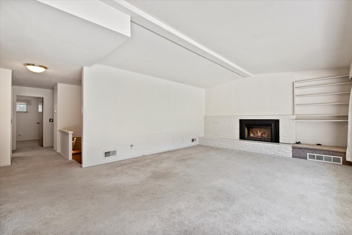 Living room with beige carpet, white walls, a white brick fireplace with a black firebox, and built-in white shelves to the right of the fireplace, ceiling with a slight drop, and an open doorway leading to a hallway.