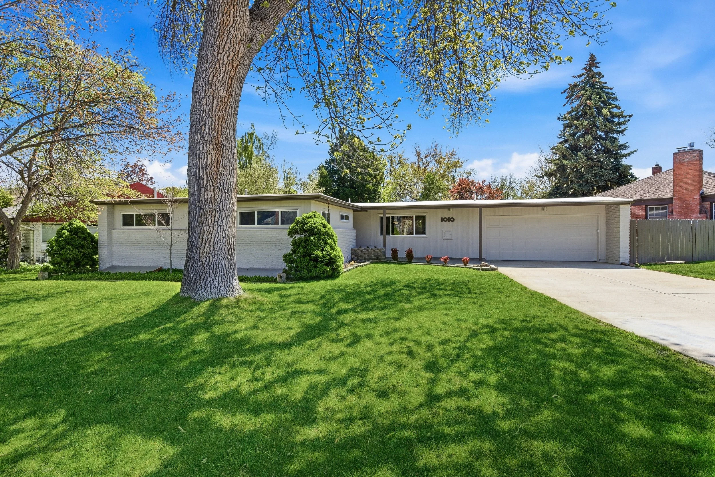 A mid-century modern house with a white exterior, flat roof, and large windows, surrounded by a well-maintained lawn with trees and bushes, under a bright blue sky with some clouds.