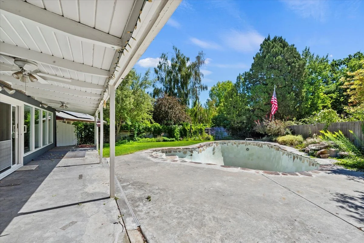 Empty backyard swimming pool with patio and lush green trees and bushes, American flag waving in the breeze, under a blue sky with some clouds.