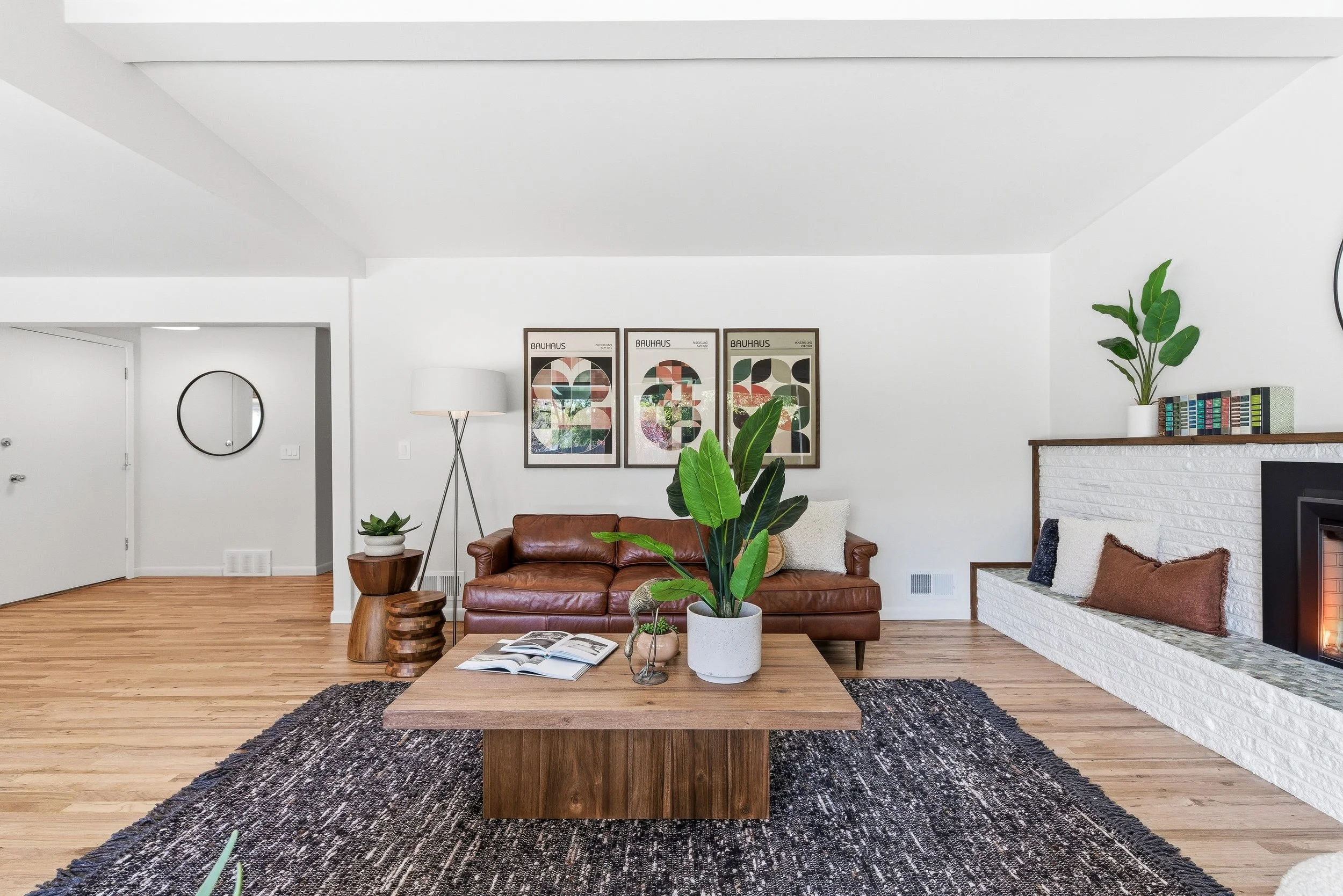 Living room with a wooden coffee table, a brown leather sofa, green potted plants, wall art, and a fireplace with white brick surround.