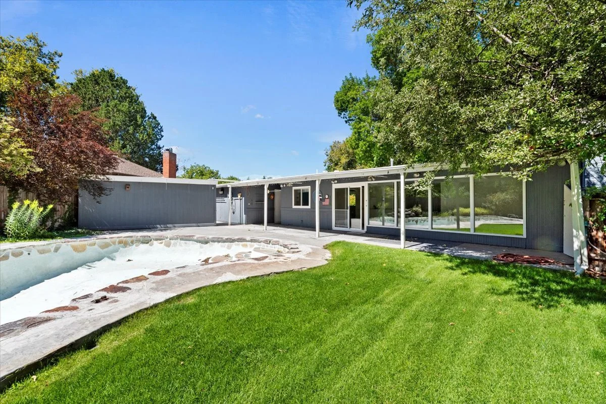 A mid-century modern house with large glass windows and sliding doors, a green lawn, and a backyard with a small stone-lined pool under a clear blue sky.